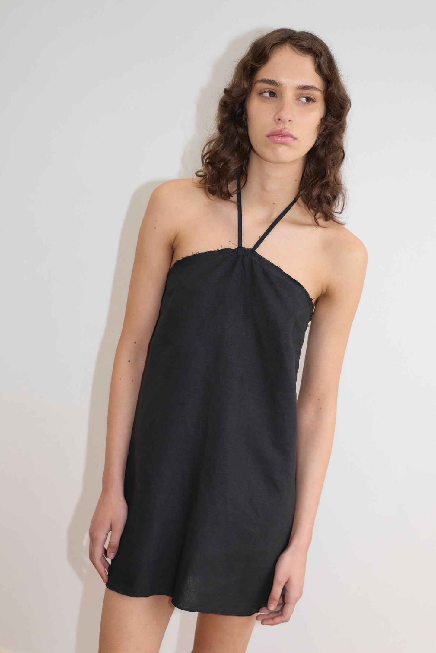 A person with wavy brown hair wears the Cross Over Dress - Black by Deiji Studios, a sleeveless halter-neck mini dress, standing against a plain white background and gazing slightly to the side with a neutral expression.