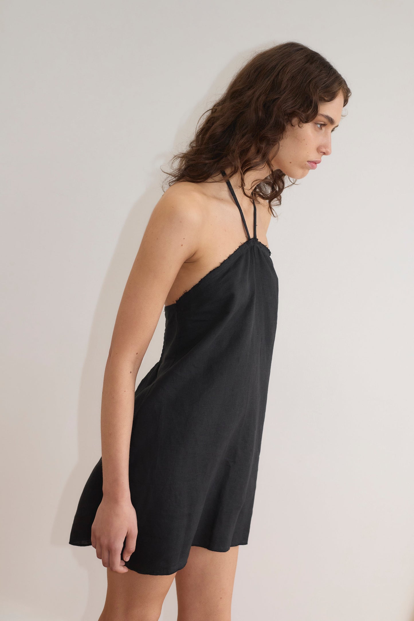 A person with wavy brown hair, in profile against a plain white background, wears a sleeveless black Cross Over Dress - Black by Deiji Studios.