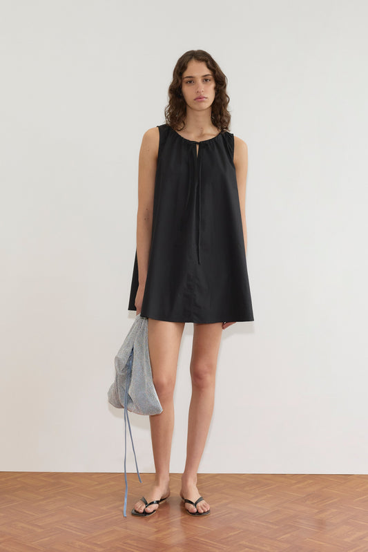 A model wears the Drawcord Dress - Black by Deiji Studios, an A-line sleeveless mini, paired with black flip-flops and a sheer silver mesh bag featuring a blue drawstring.