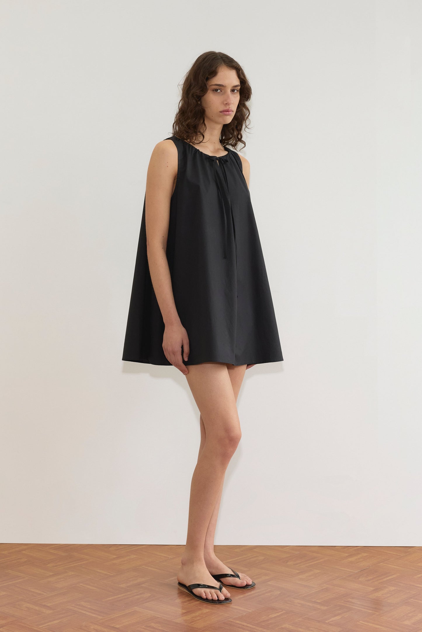 A woman with wavy brown hair stands indoors against a white wall, wearing the Deiji Studios Drawcord Dress - Black and black flip-flops. She looks slightly to the side with a neutral expression.
