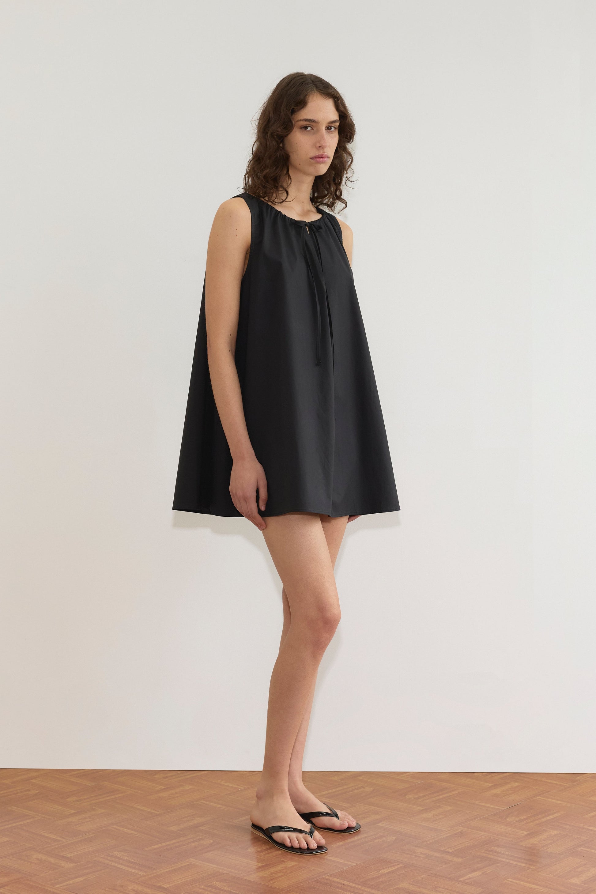 A woman with wavy brown hair stands indoors against a white wall, wearing the Deiji Studios Drawcord Dress - Black and black flip-flops. She looks slightly to the side with a neutral expression.