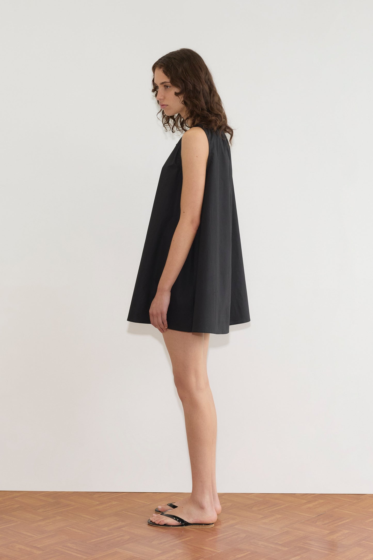 A person with shoulder-length curly hair stands sideways on a wooden floor, wearing the loose-fitting Drawcord Dress - Black by Deiji Studios and black sandals against a plain white wall.