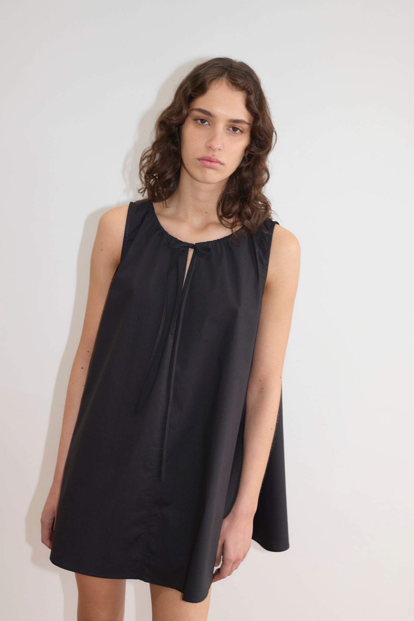 A woman with wavy brown hair wears the Drawcord Dress - Black, a sleeveless, loose-fit dress featuring a small front keyhole tie and subtle A-line shape, standing against a plain white background.