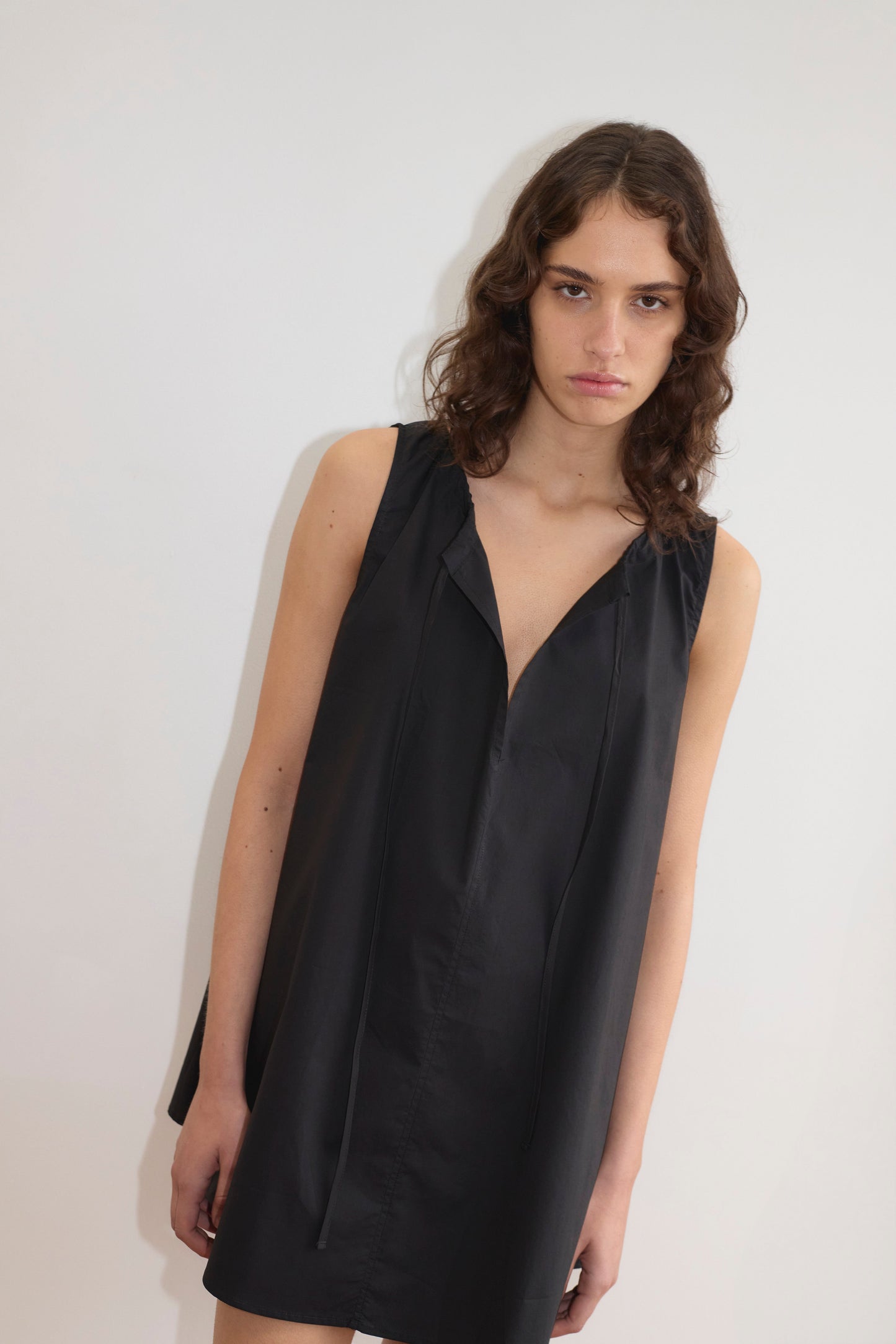 A person with wavy brown hair wears Deiji Studios' Drawcord Dress - Black, a sleeveless, loose-fitting a-line dress, standing against a plain white background and looking at the camera with a neutral expression.