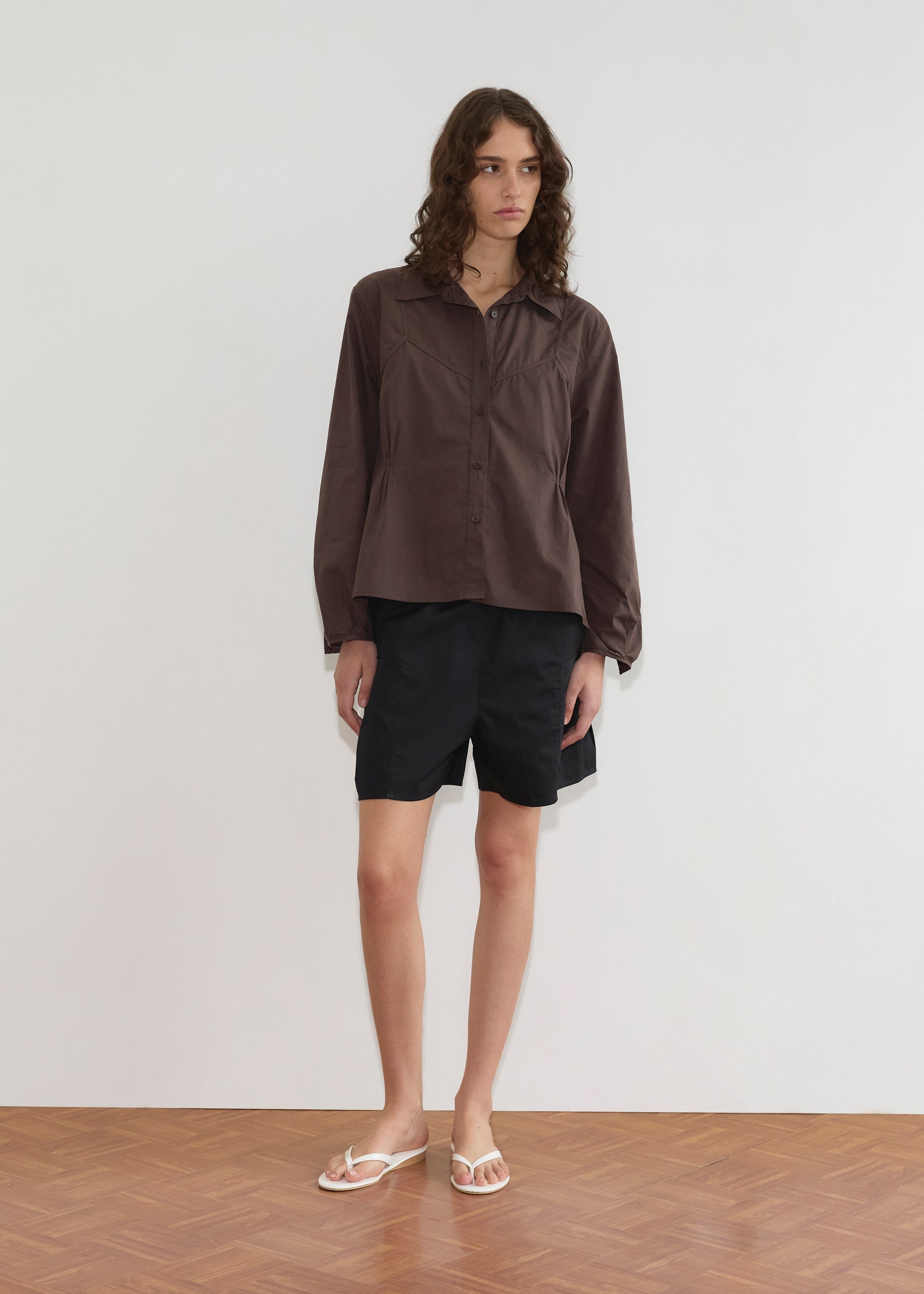 A person with wavy brown hair stands indoors against a white wall, wearing a loose brown Deiji Studios button-up shirt, Pleat Short - Black, and white flip-flops on a wooden floor.