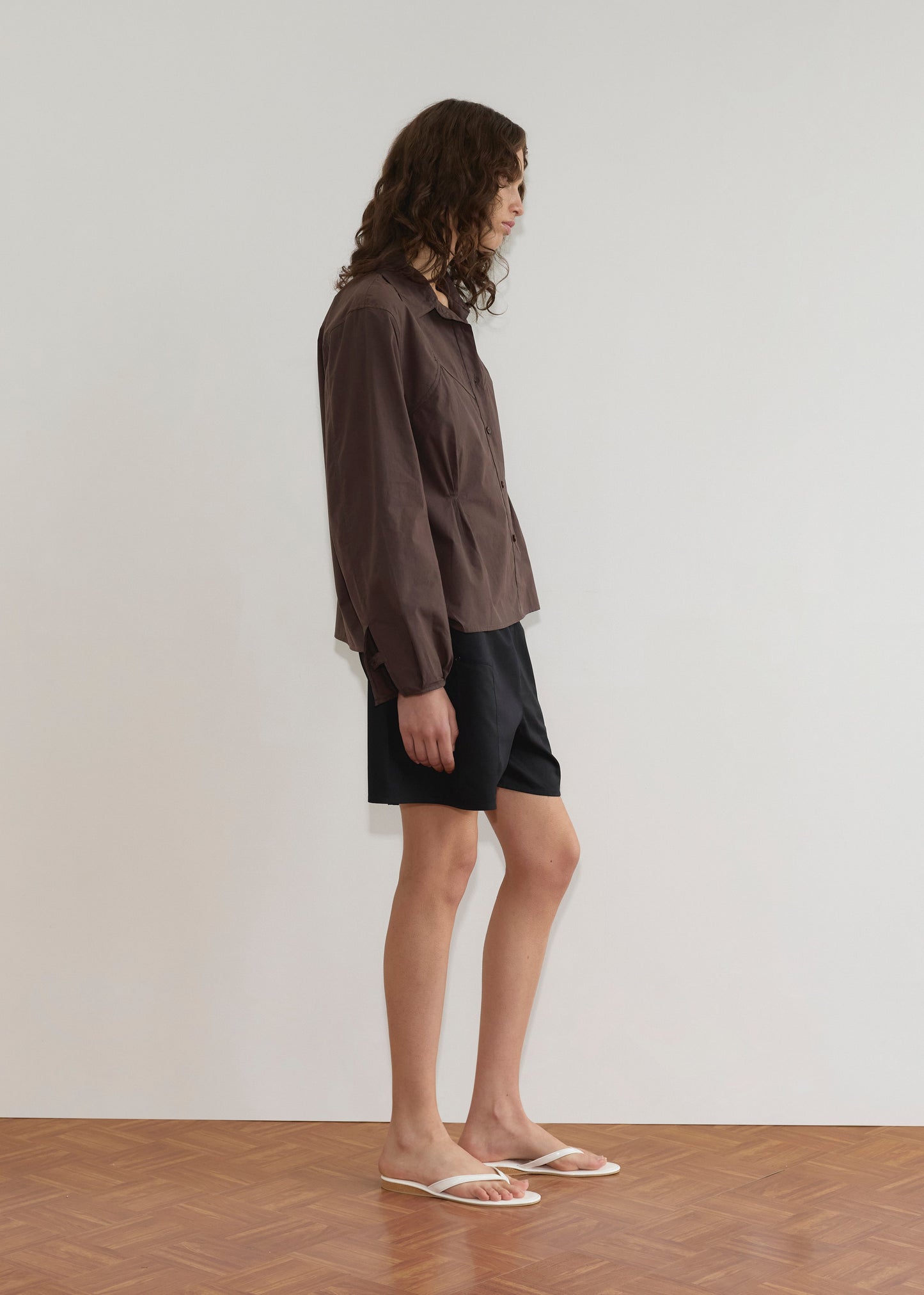 A person with long, wavy hair stands sideways wearing a brown button-up shirt, Pleat Short - Black by Deiji Studios, and white flip-flops against a plain white background on a wooden floor.