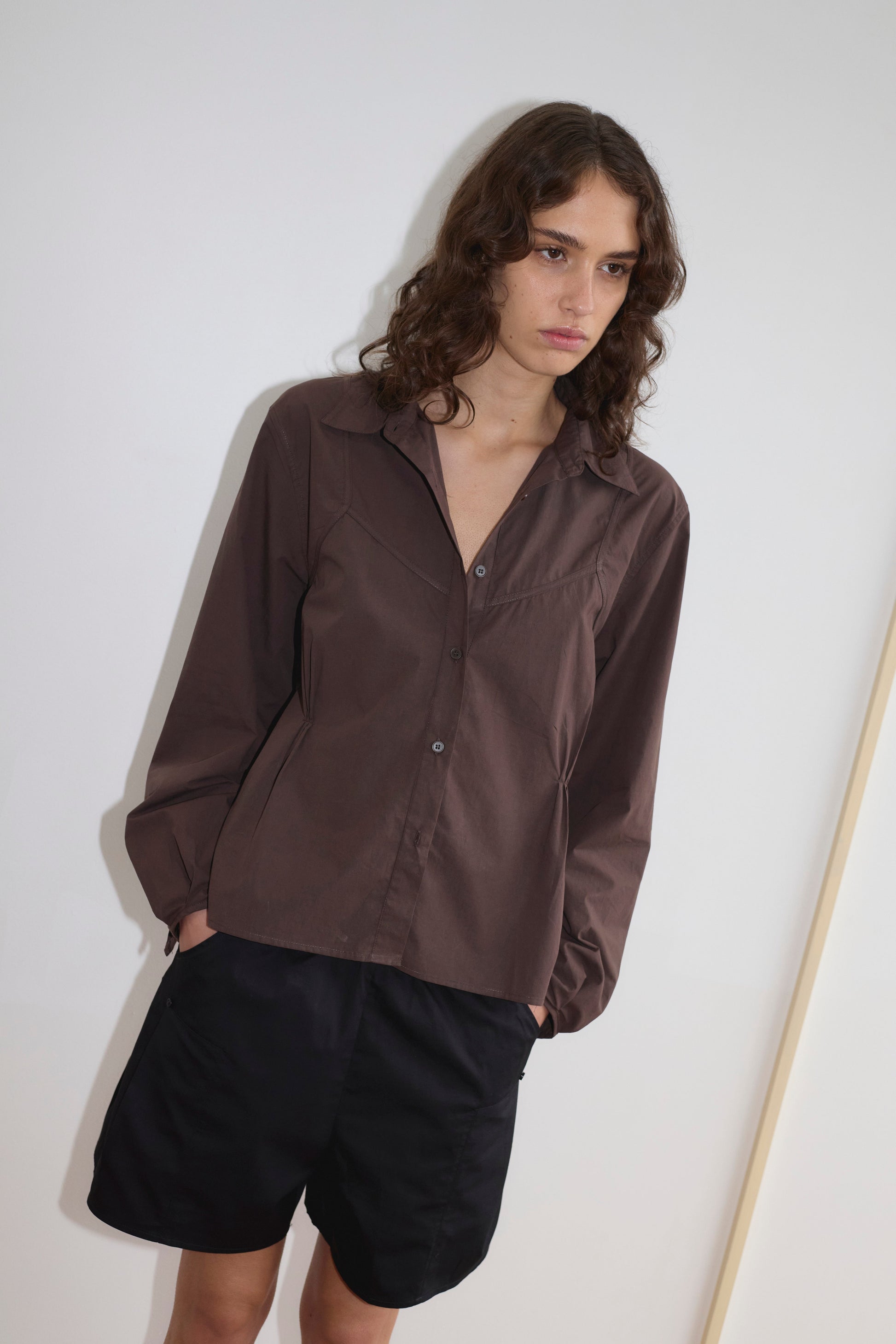 A person with wavy brown hair wears a brown Deiji Studios button-up shirt and relaxed black Pleat Shorts, standing against a plain white wall with hands in their pockets, looking slightly downward with a neutral expression.