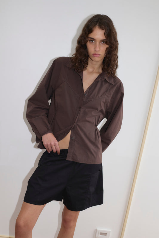 A person with long wavy hair wears a brown button-up shirt and the Pleat Short - Black from Deiji Studios, standing against a plain white wall with a neutral expression.