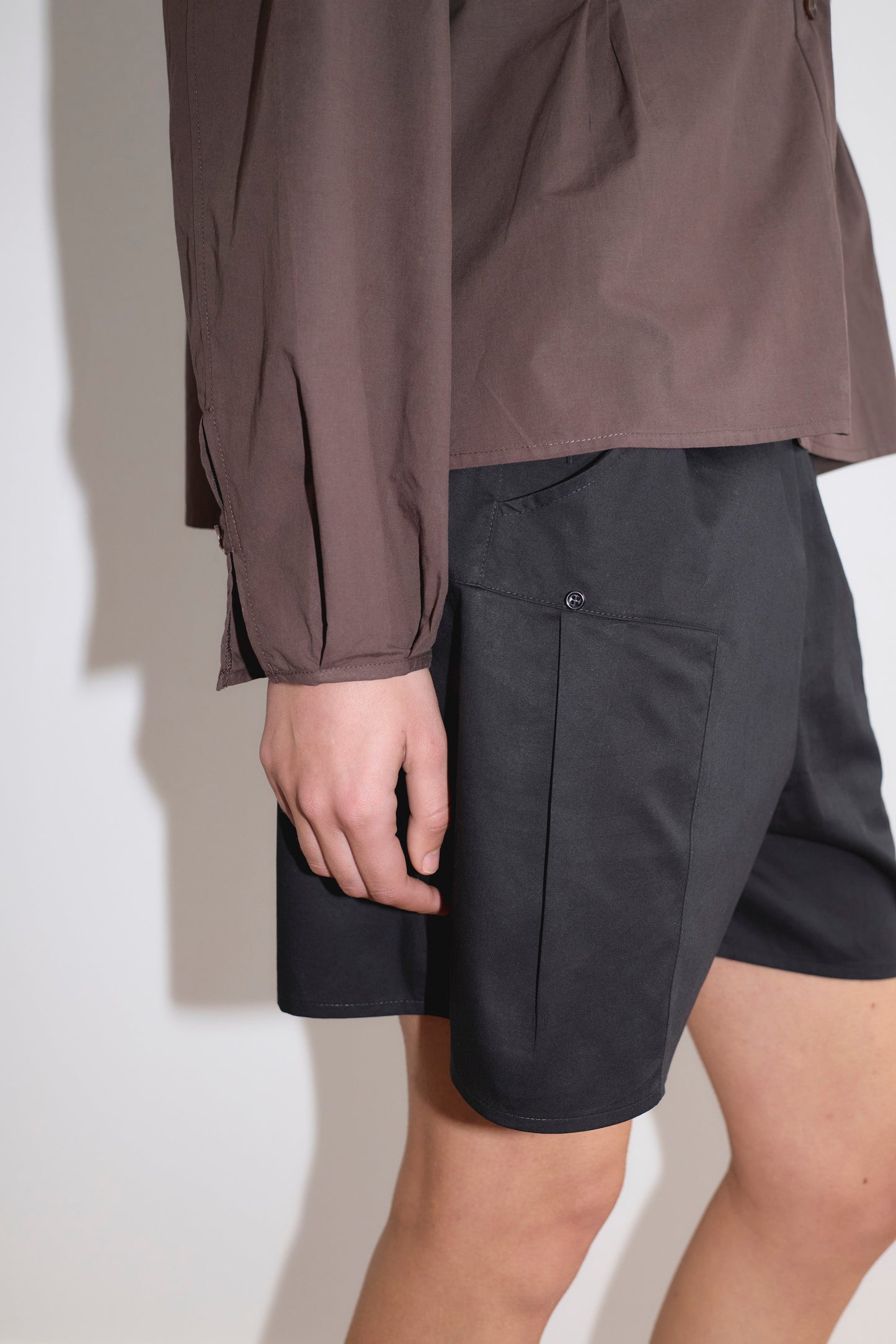 Wearing a long-sleeved brown shirt with Pleat Short - Black, the person stands against a plain light background, showing only the lower torso, arm, and upper legs.