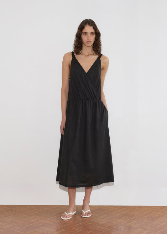 A woman with wavy brown hair wears the Crossed Dress, a sleeveless black wrap dress at knee length, paired with white sandals. She stands against a plain white wall on a wooden floor.