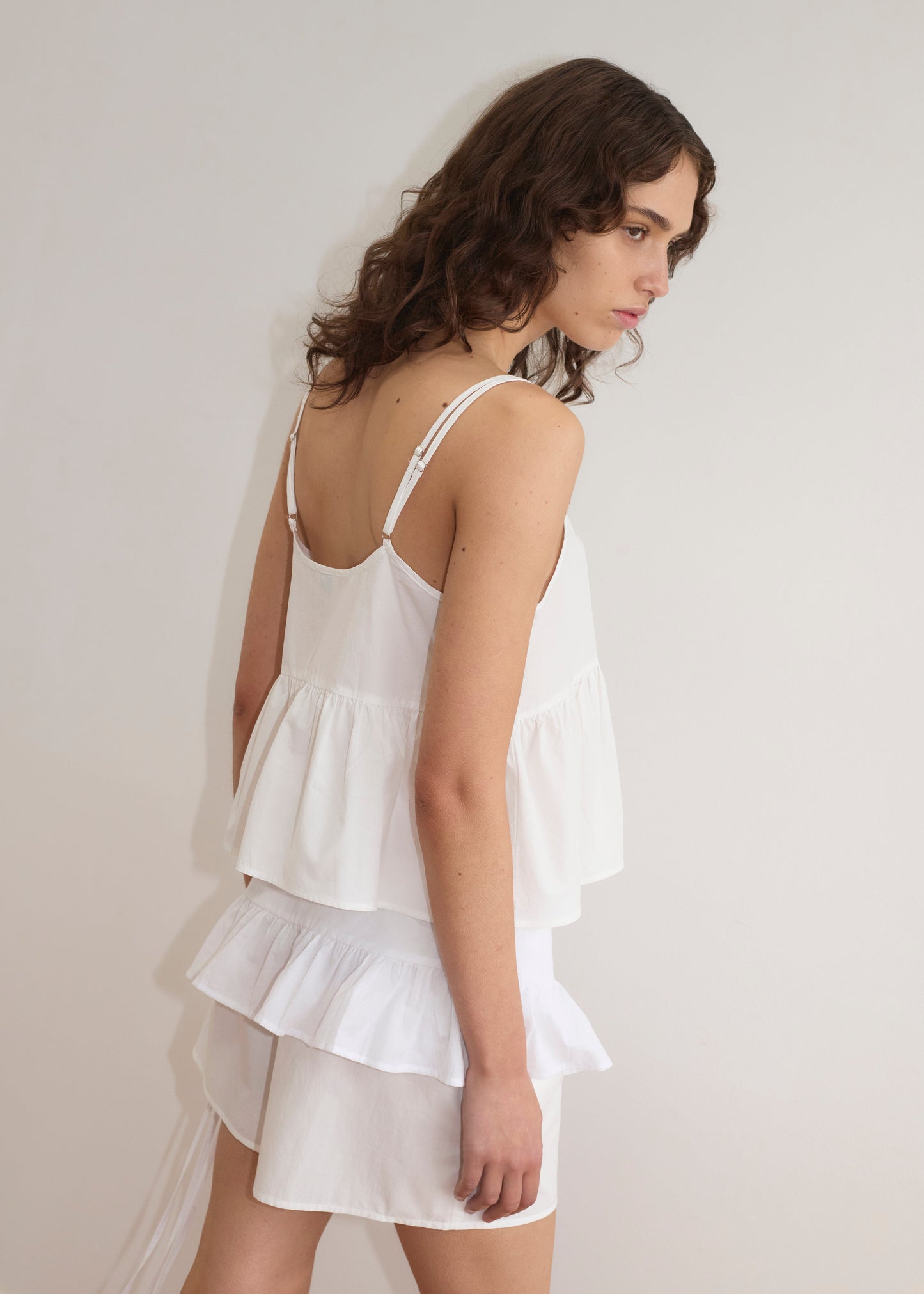 A woman with wavy brown hair stands sideways against a plain white wall, wearing the Flounce Belt - White as a sleeveless ruffled top with adjustable ties and paired with a matching skirt.