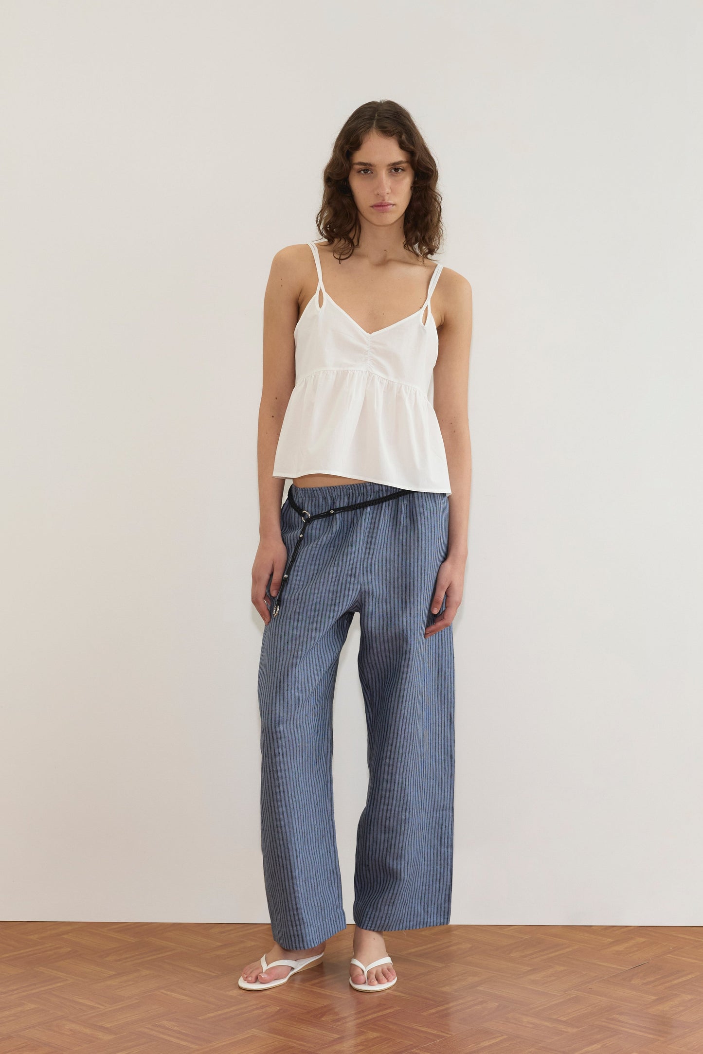 A person wearing the Bind Top - White by Deiji Studios, blue striped wide-leg pants with a braided belt, and white sandals stands against a plain white wall on a wooden floor.