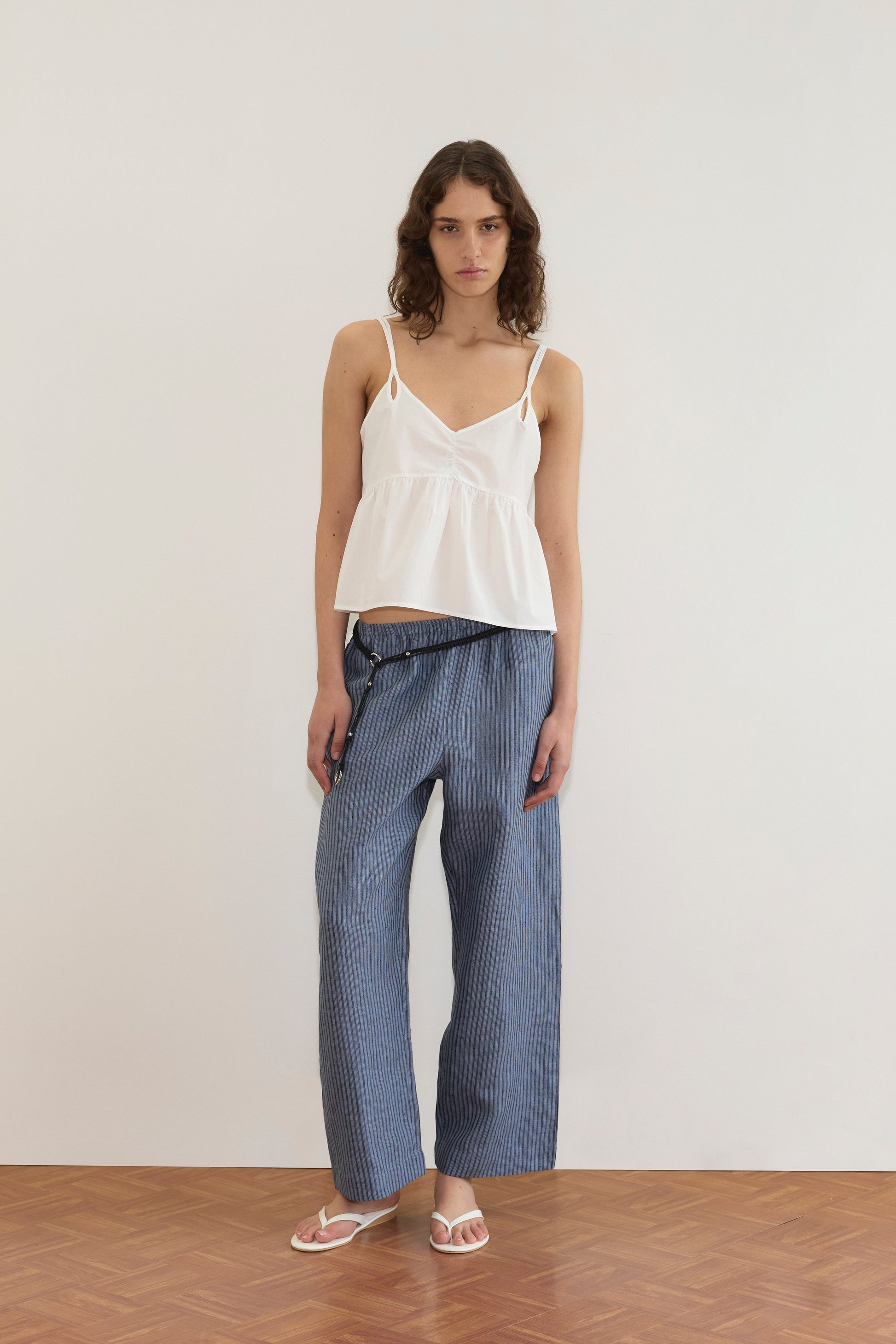 A woman stands against a white wall wearing a sleeveless white top, Linen Ease Trouser in Azure Stripe from Deiji Studios, white sandals, and a thin black belt. Her hair is down in loose waves.