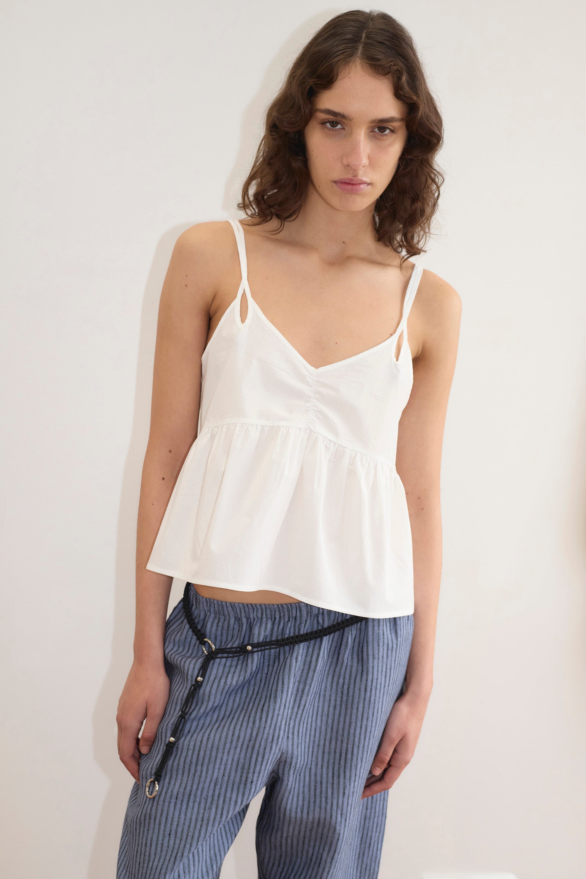 A woman with wavy brown hair wears the Bind Top - White from Deiji Studios and blue pinstriped pants, standing against a plain white wall with a relaxed pose and neutral expression.