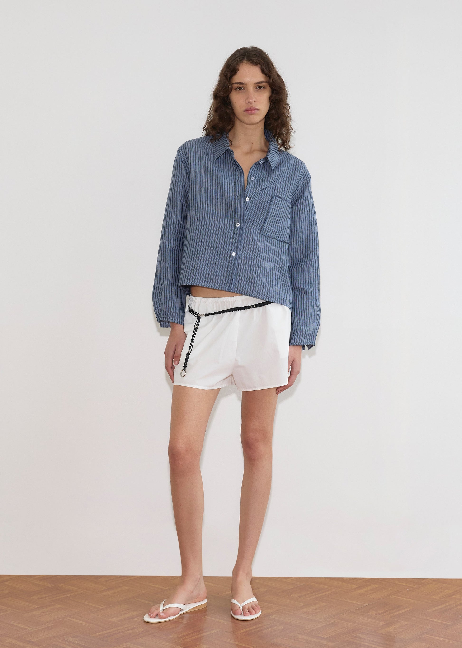A person stands indoors against a plain white wall, wearing the Double Placket Shirt - Azure Stripe, white shorts with a black zipper detail, and white flip-flops. They have long curly hair and a neutral expression.