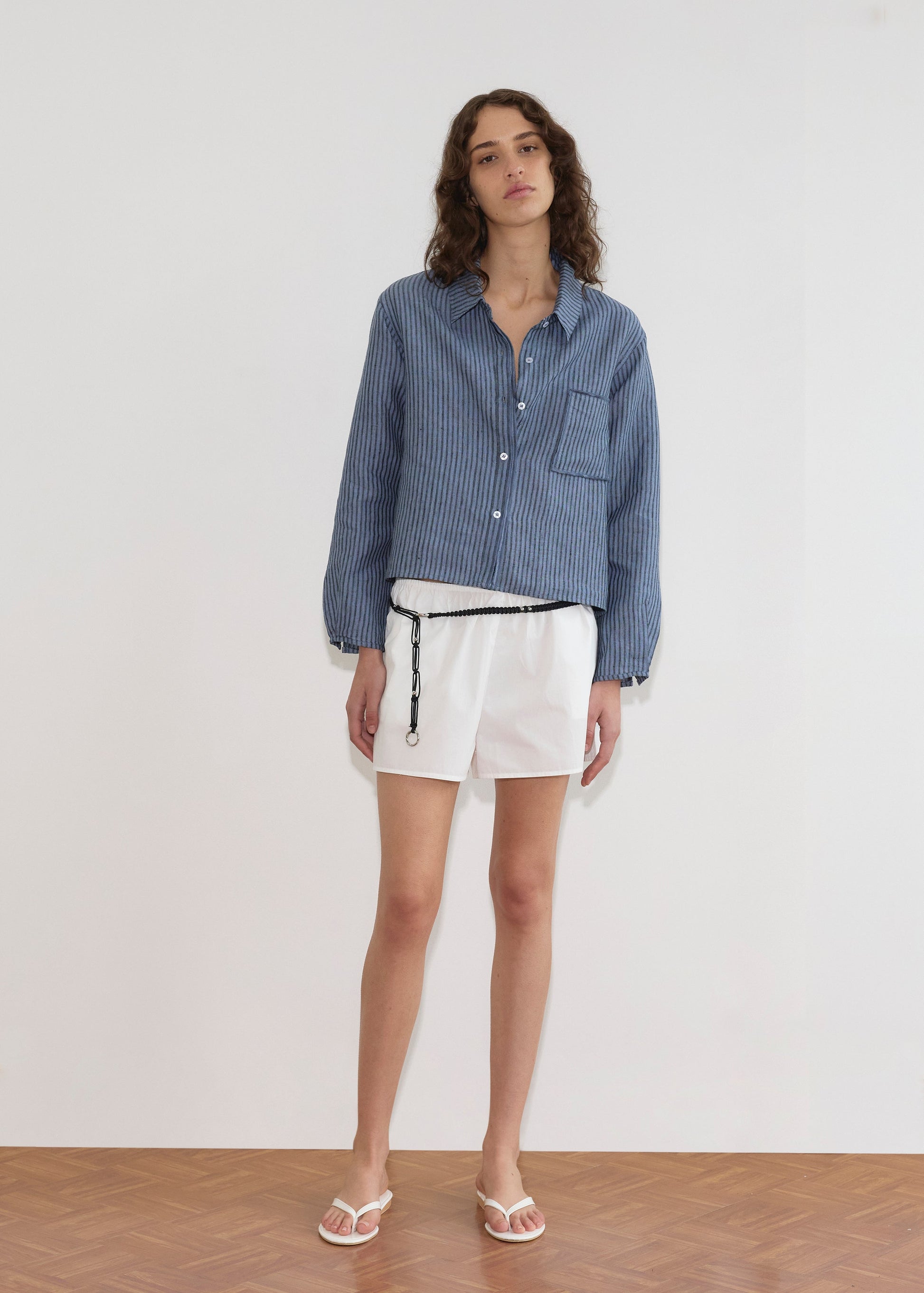 A woman with wavy brown hair and a relaxed expression stands against a plain white wall wearing the Double Placket Shirt - Azure Stripe, paired with white shorts, a black belt, and white sandals.