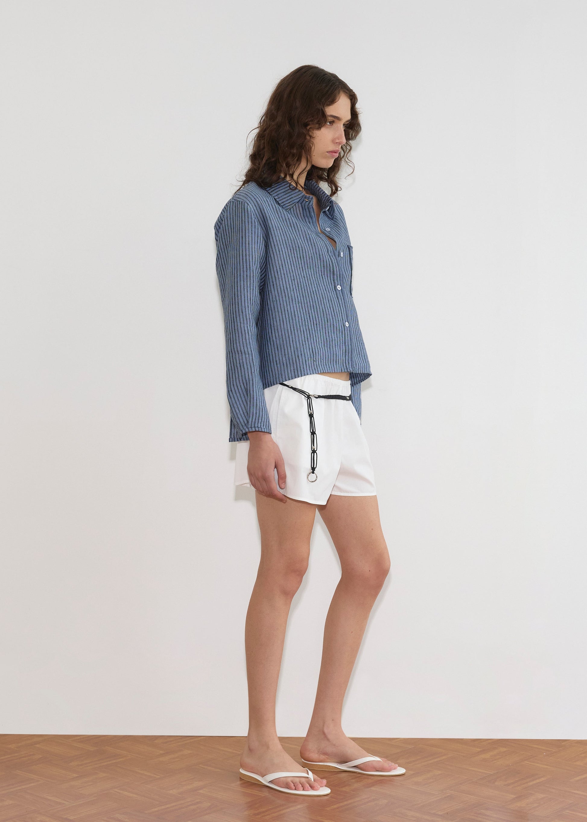A person with wavy brown hair wears the Double Placket Shirt - Azure Stripe, paired with white shorts featuring a black chain and white flip-flops, standing indoors on a wooden floor against a plain white wall.