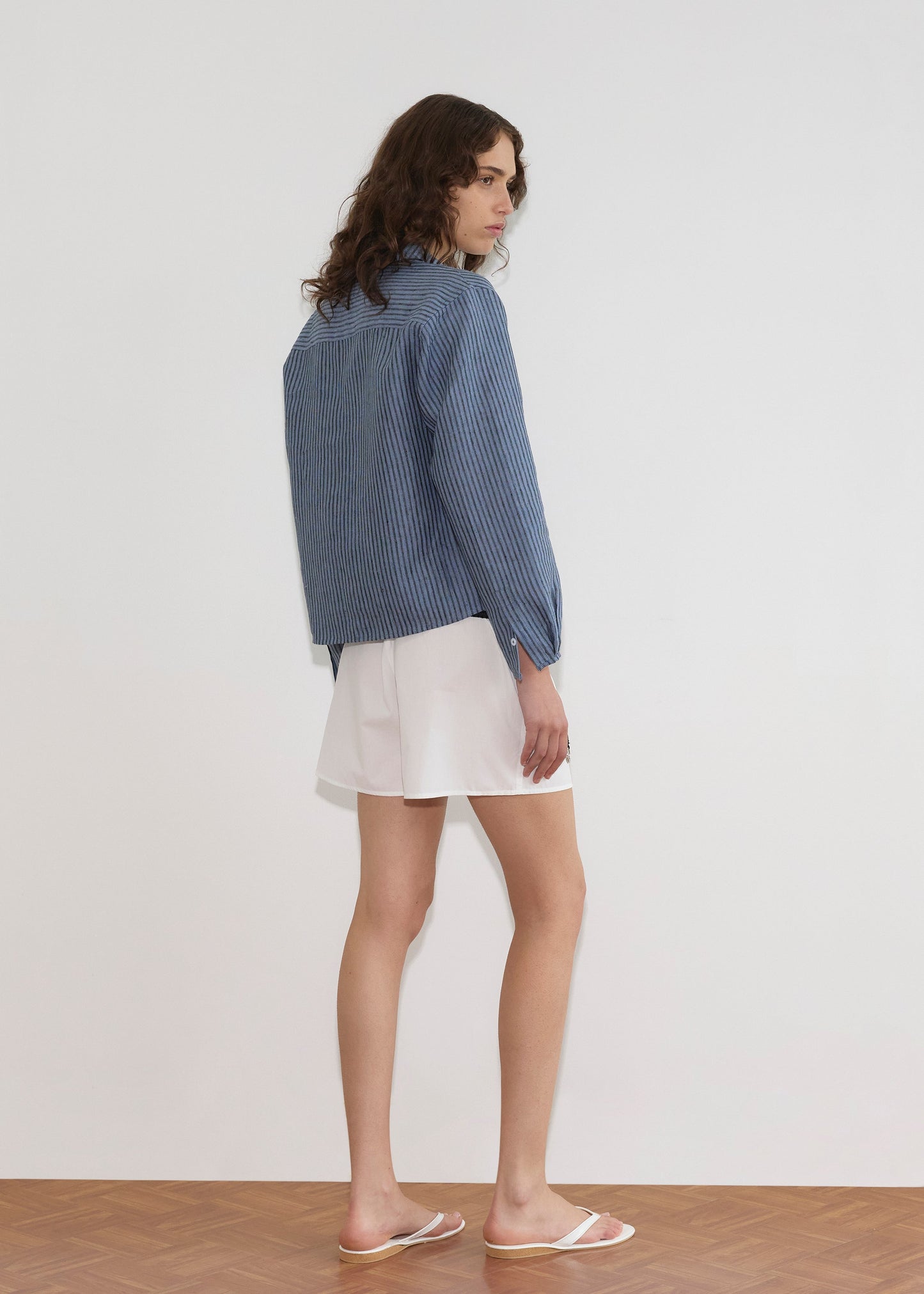 A person with wavy brown hair stands on a wooden floor, facing away. They wear the Double Placket Shirt - Azure Stripe with white shorts and sandals against a plain white wall.