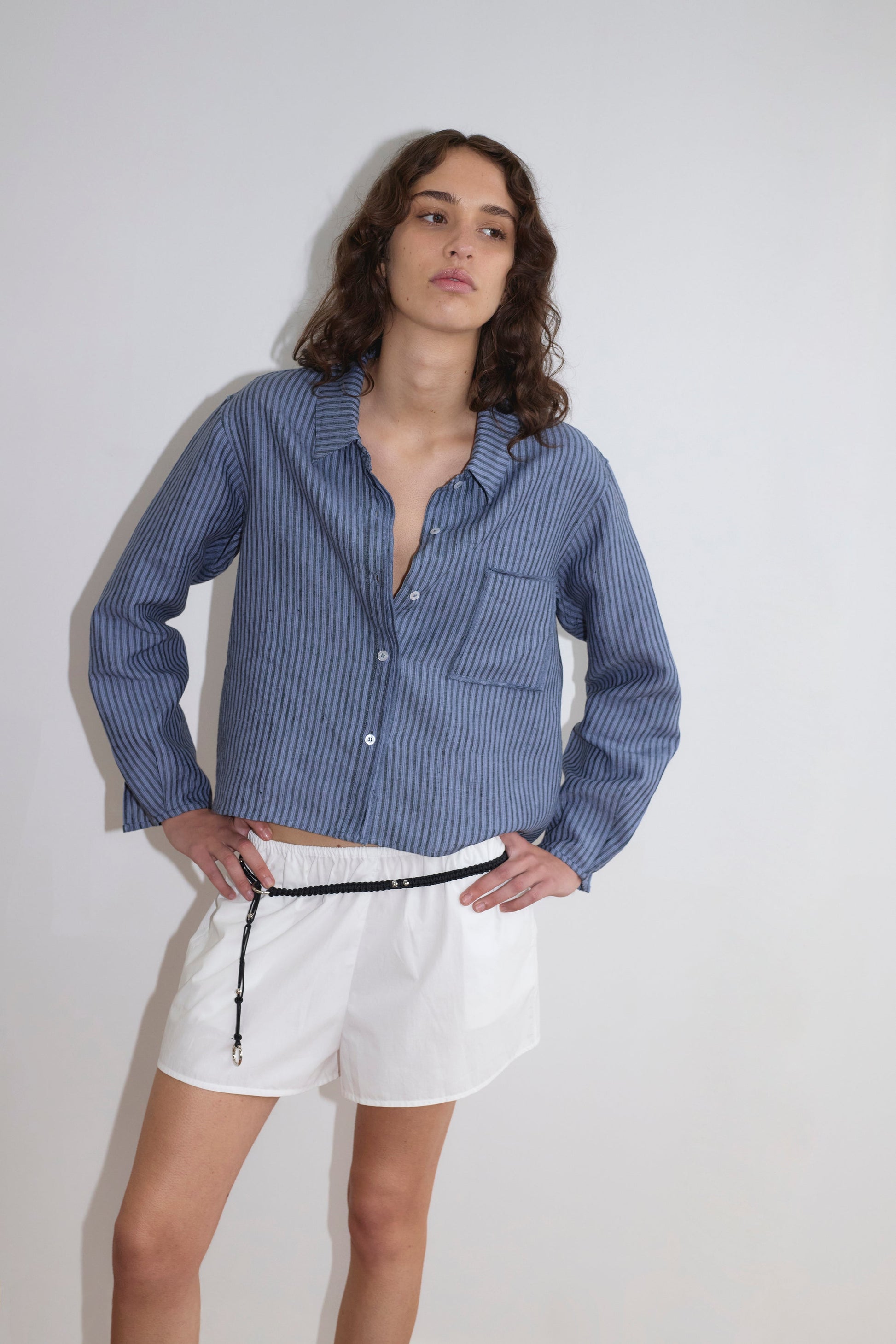 A person poses against a white background wearing the Double Placket Shirt - Azure Stripe, styled with white shorts and a black cord belt, one hand on their hip.