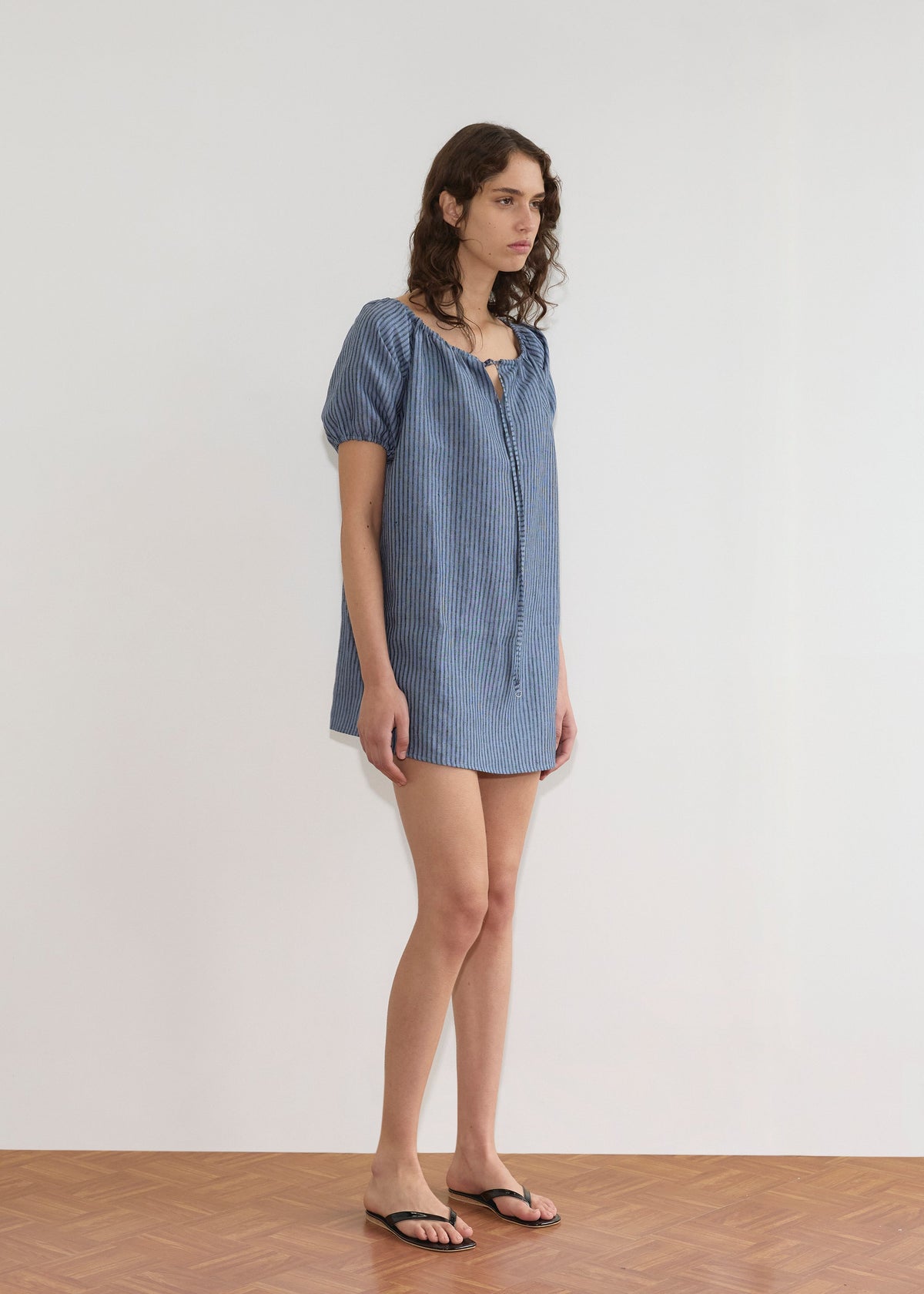 A person with wavy brown hair stands indoors on a wooden floor, wearing the Smock Dress - Azure Stripe by Deiji Studios and black flip-flops, facing slightly to the side against a plain white wall.