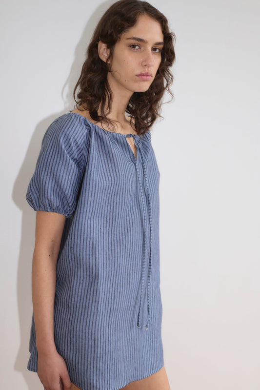 A person with wavy brown hair wears the Smock Dress - Azure Stripe from Deiji Studios, a loose-fitting blue striped short-sleeve dress, and stands against a plain white background, gazing slightly to the side with a neutral expression.