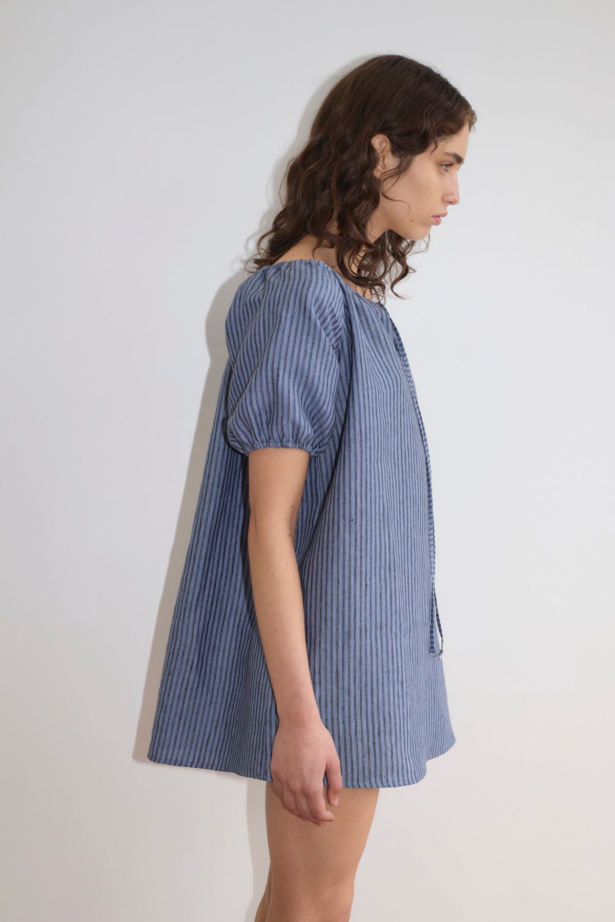 A person with wavy brown hair stands sideways against a plain light wall, wearing the Smock Dress - Azure Stripe from Deiji Studios, featuring blue and gray vertical stripes and puffed short sleeves.