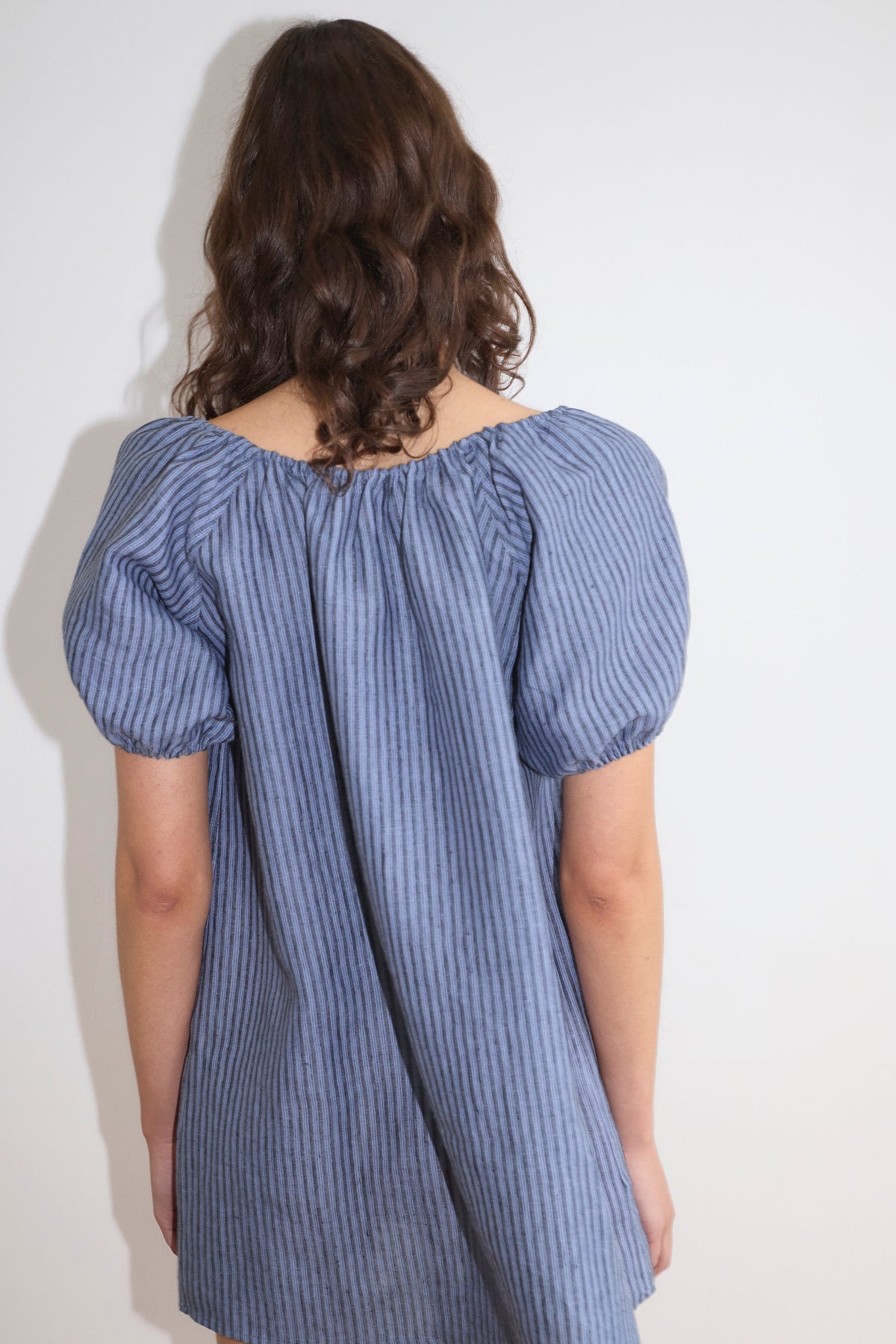 A person with wavy brown hair stands with their back to the camera, wearing the Smock Dress - Azure Stripe with short sleeves, against a plain white background.