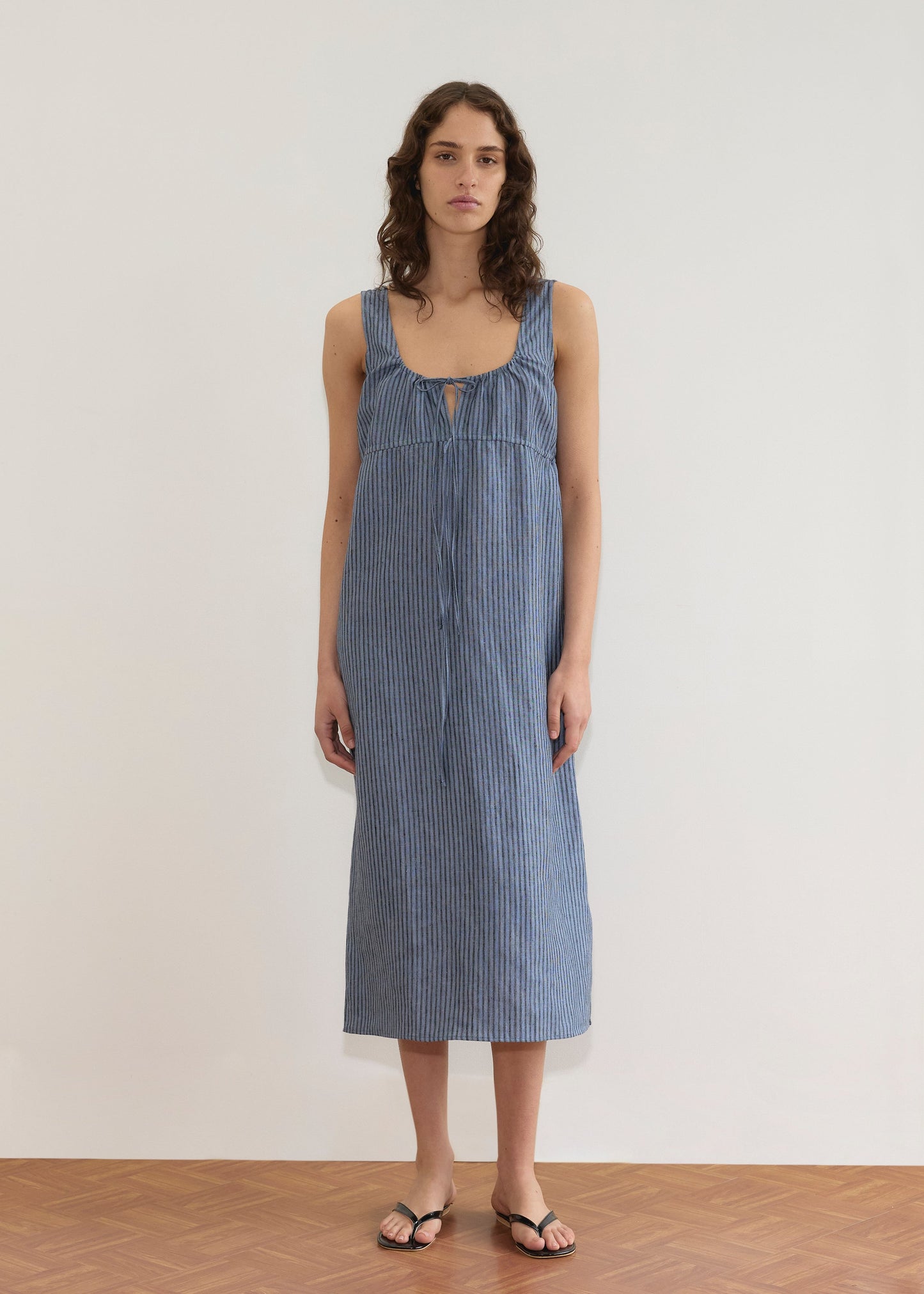 A woman stands against a plain white wall wearing the Sleeveless Cord Dress - Azure Stripe, an ankle-length, blue and white striped midi dress with a front tie detail, paired with black sandals on a wooden floor.
