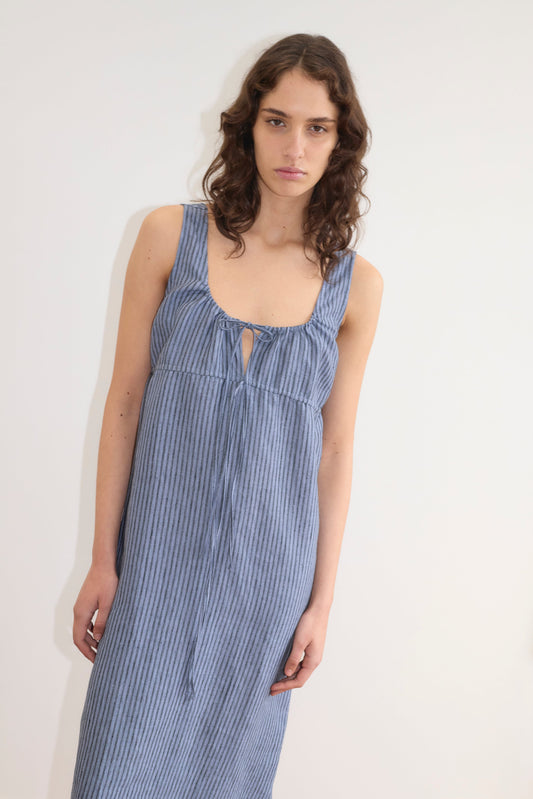 A woman with wavy brown hair wears the Deiji Studios Sleeveless Cord Dress - Azure Stripe, featuring blue and white stripes, a scoop neckline, and front tie detail, standing against a plain white background.