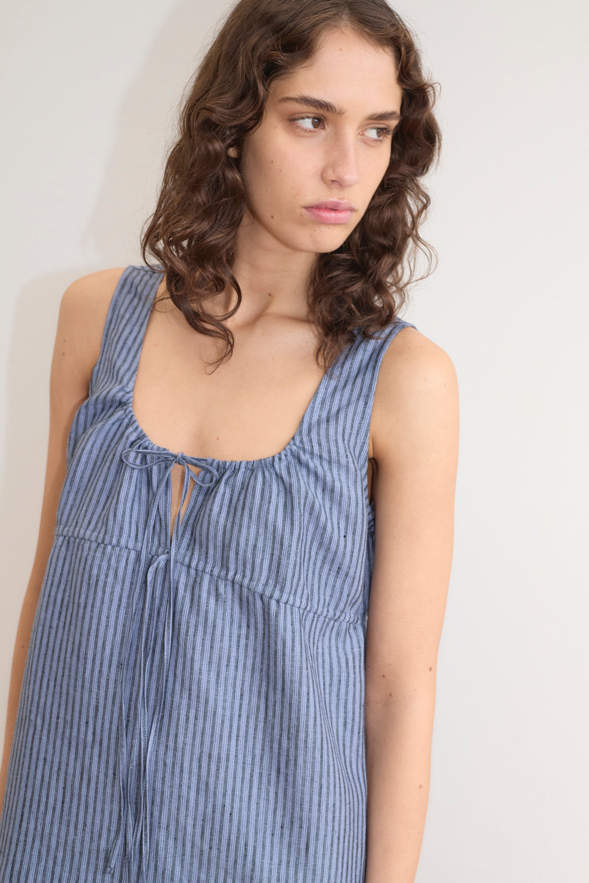 A person with wavy brown hair wears the Deiji Studios Sleeveless Cord Dress - Azure Stripe, featuring blue and gray stripes, a scoop neckline, and bow detail, looking to the side against a plain light background.