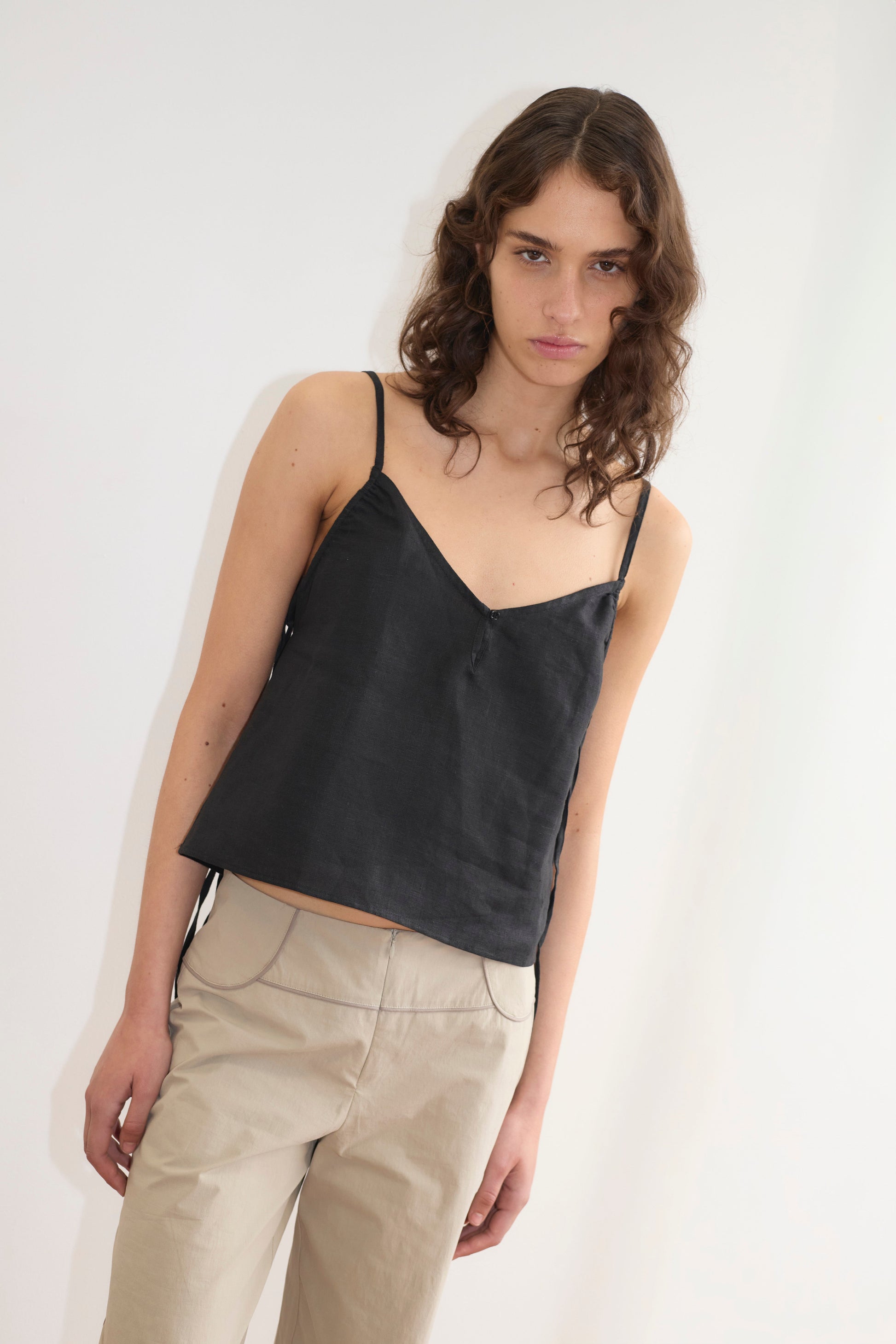 A model with curly brown hair wears the V Neck Cami in black satin with spaghetti straps and a V-neckline, paired with beige wide-leg trousers, against a white background.