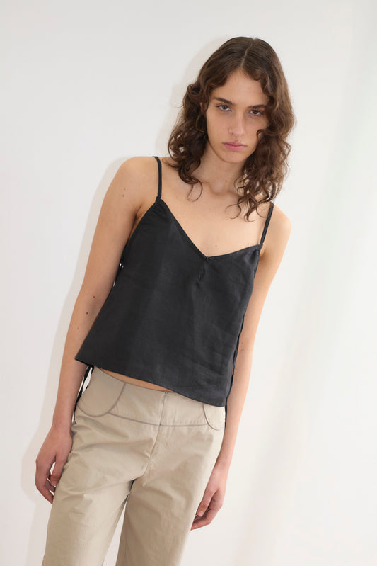 A model with curly brown hair wears the V Neck Cami in black satin with spaghetti straps and a V-neckline, paired with beige wide-leg trousers, against a white background.