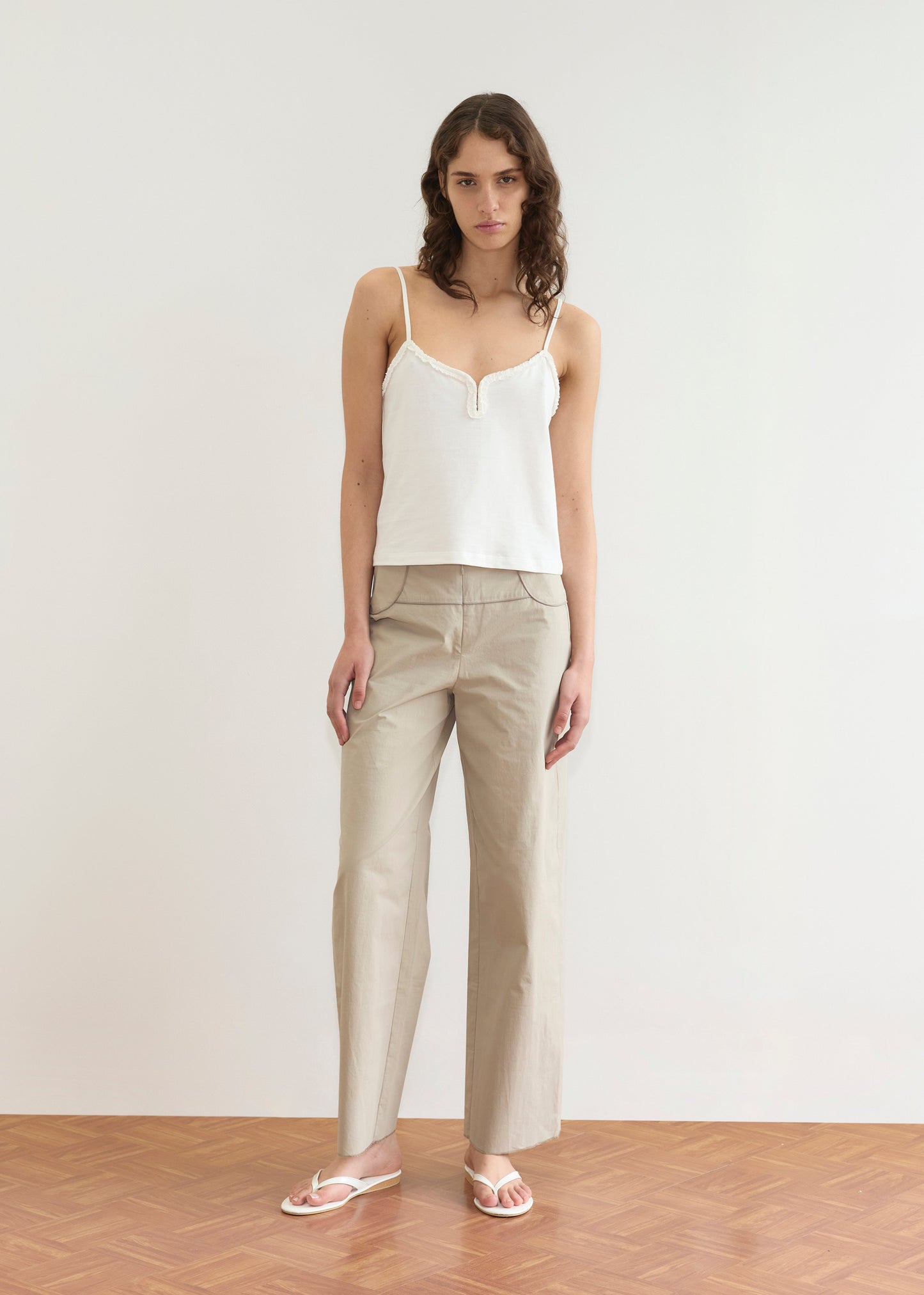 A woman with long wavy hair stands indoors on a wooden floor against a white wall, wearing the Clasp Singlet - White by Deiji Studios with adjustable straps, beige wide-leg trousers, and white sandals. She has a neutral expression.