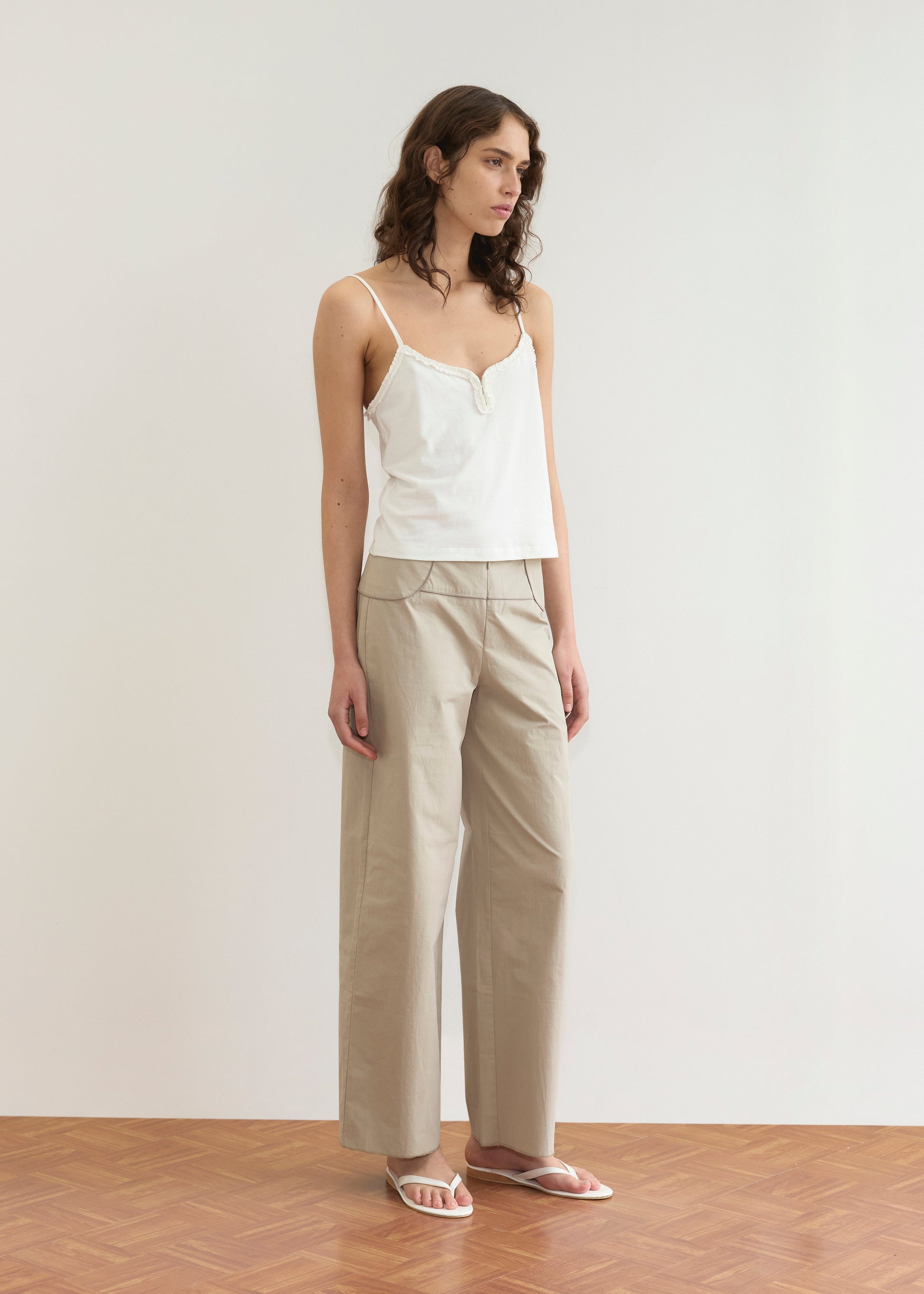 A woman stands indoors in a white camisole top, Deiji Studios’ Panelled Pant - Stone with a straight-leg silhouette, and white sandals. She has wavy brown hair and looks to the side against a plain light background.