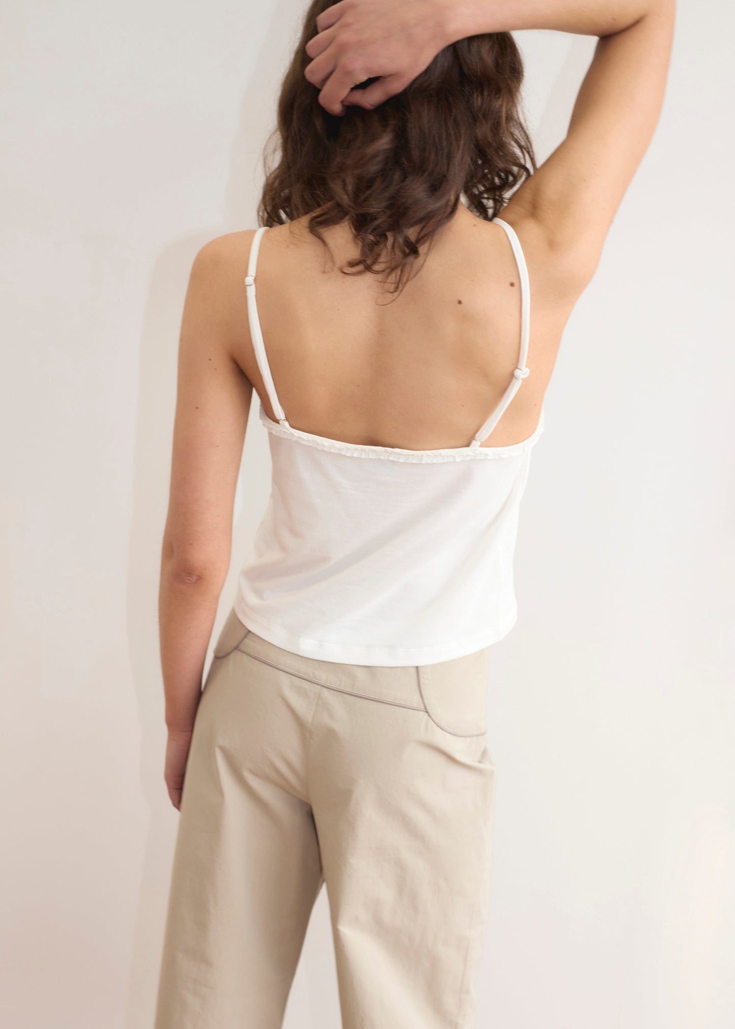 A person with wavy brown hair, wearing the Clasp Singlet - White by Deiji Studios and beige pants, stands with their back to the camera, one arm raised and hand resting on their head.