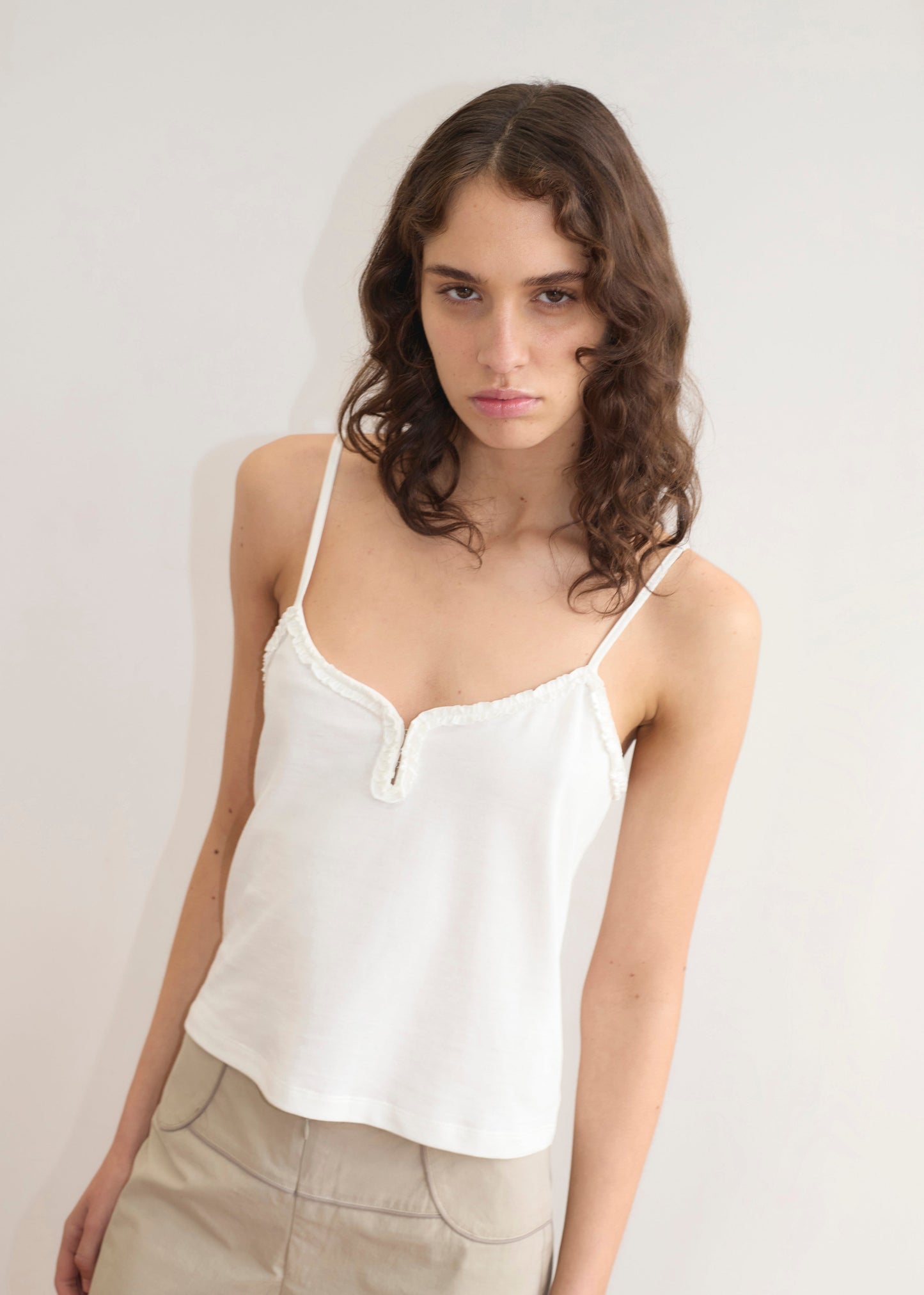 A young woman with wavy brown hair wears the Clasp Singlet - White by Deiji Studios, featuring adjustable straps, paired with beige pants, standing against a plain light-colored wall and looking calmly at the camera.
