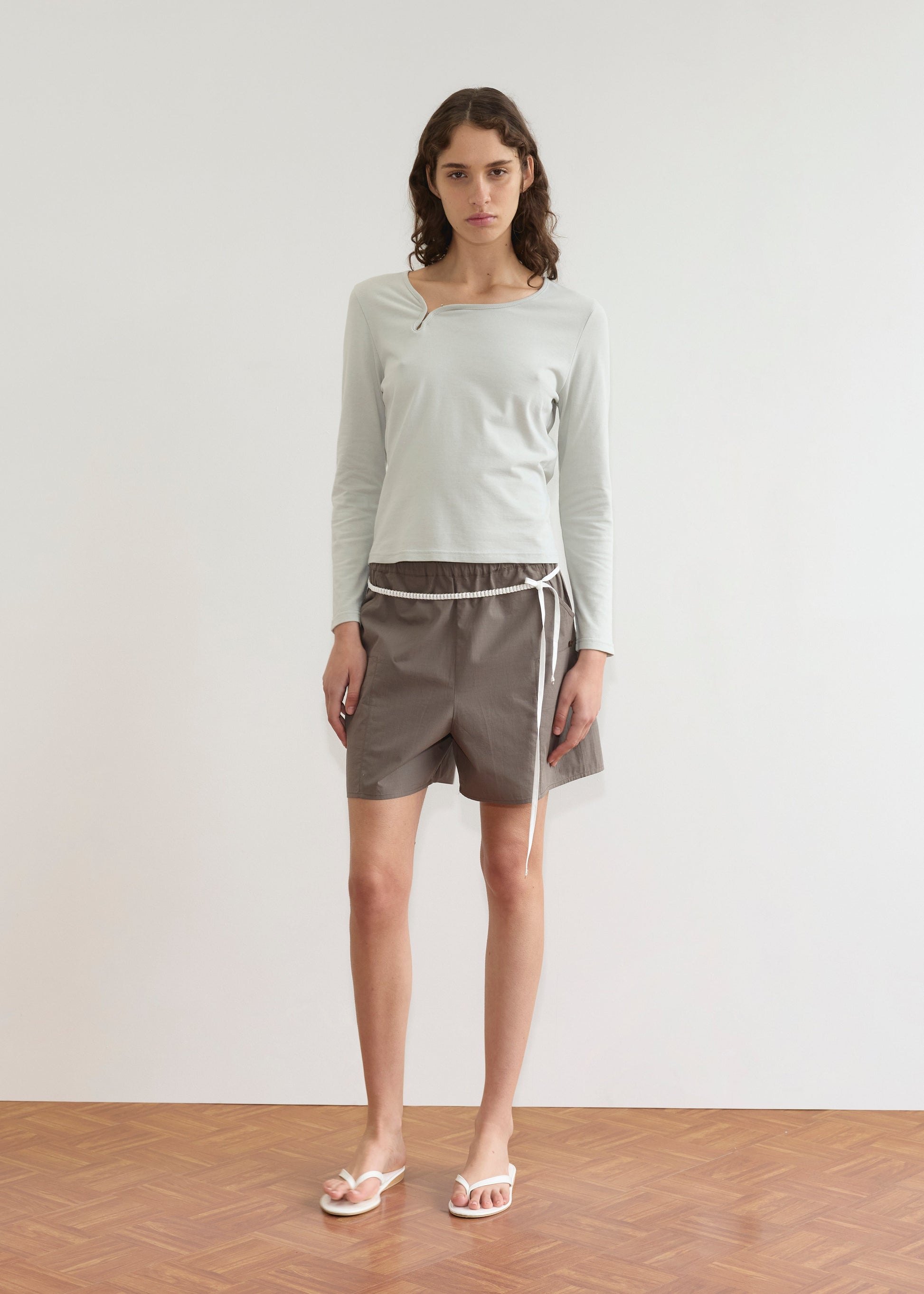 A person with long brown hair stands indoors against a plain light wall, wearing a Deiji Studios long-sleeve top, Pleat Short - Khaki Check, and white flip-flops on a wooden floor.