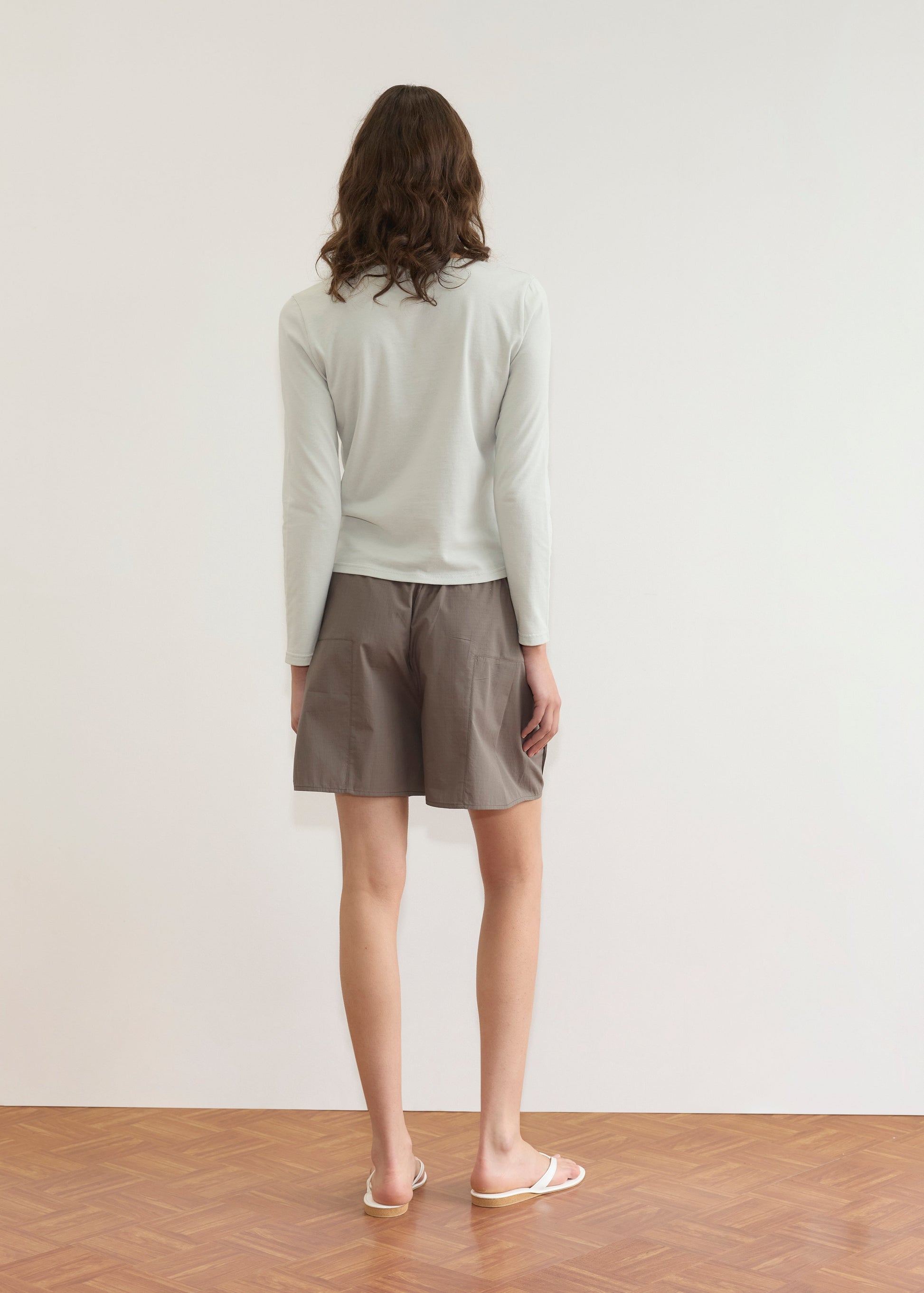 A person with shoulder-length brown hair stands facing a white wall, wearing a light long-sleeve shirt, Deiji Studios Pleat Short - Khaki Check, and white flip-flops on a wooden floor.