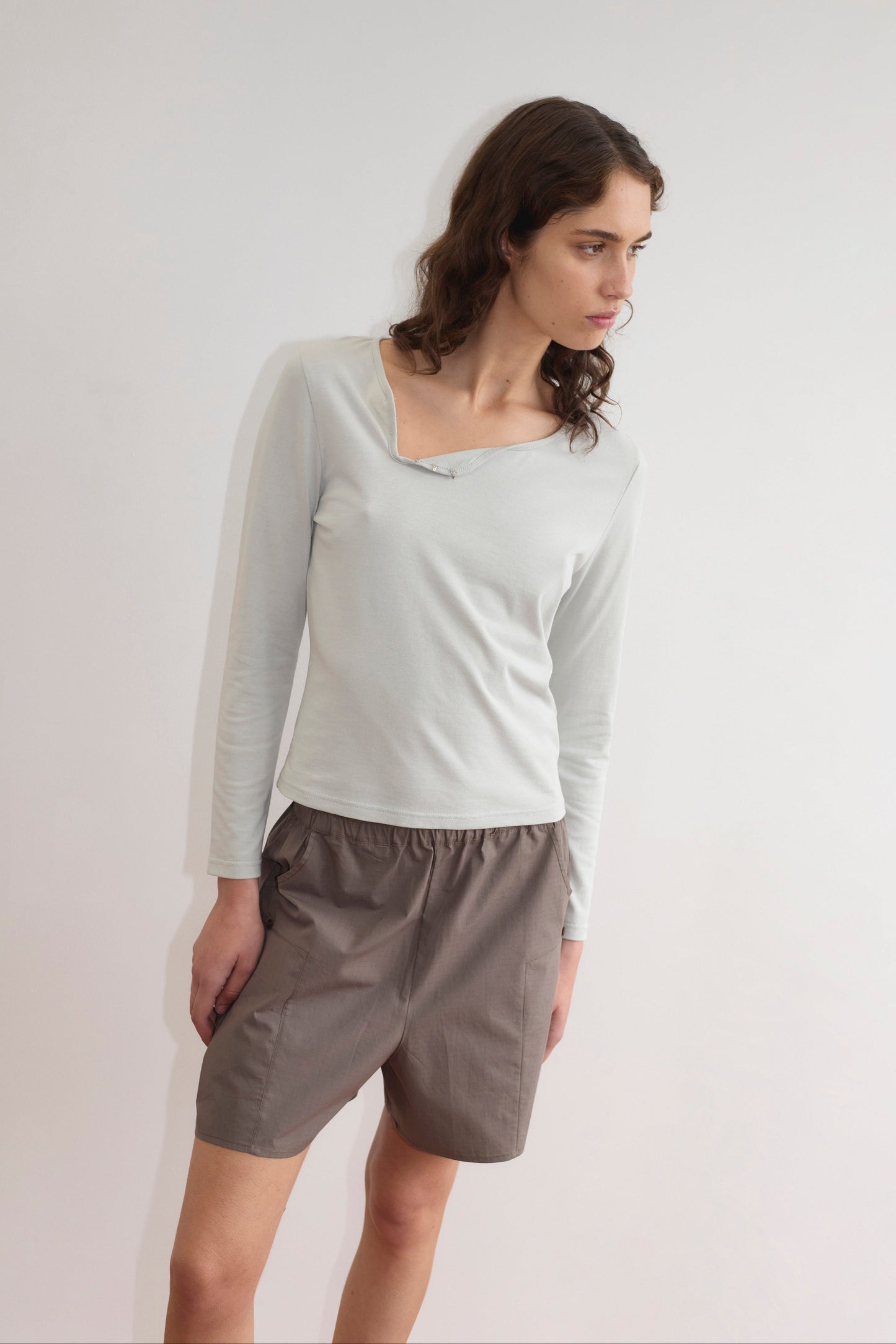 A woman with wavy brown hair wears a light long-sleeve top with an asymmetrical neckline and the Pleat Short - Khaki Check, standing against a plain white background and looking slightly to her right.