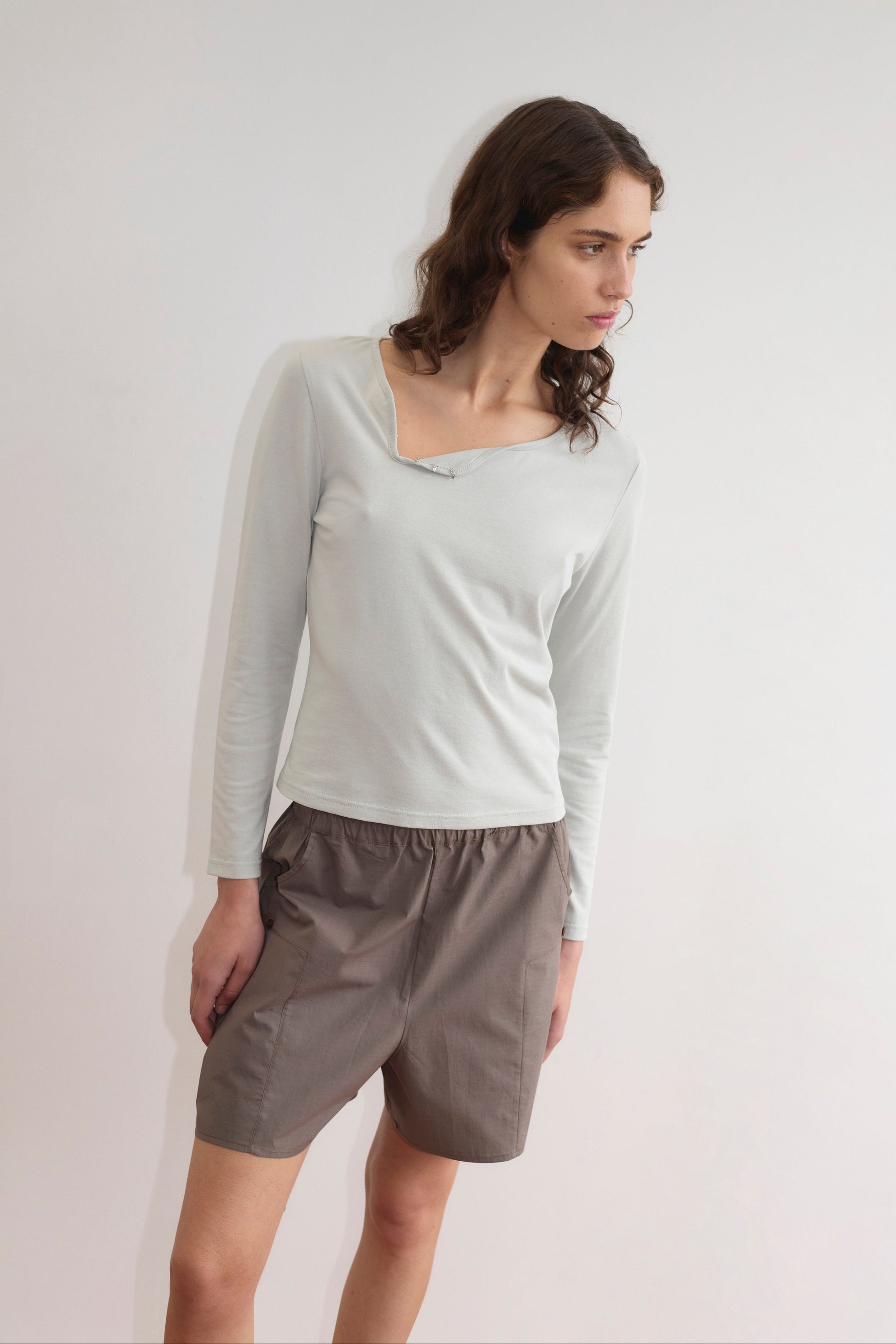 A woman with wavy brown hair wears a light long-sleeve top with an asymmetrical neckline and the Pleat Short - Khaki Check, standing against a plain white background and looking slightly to her right.