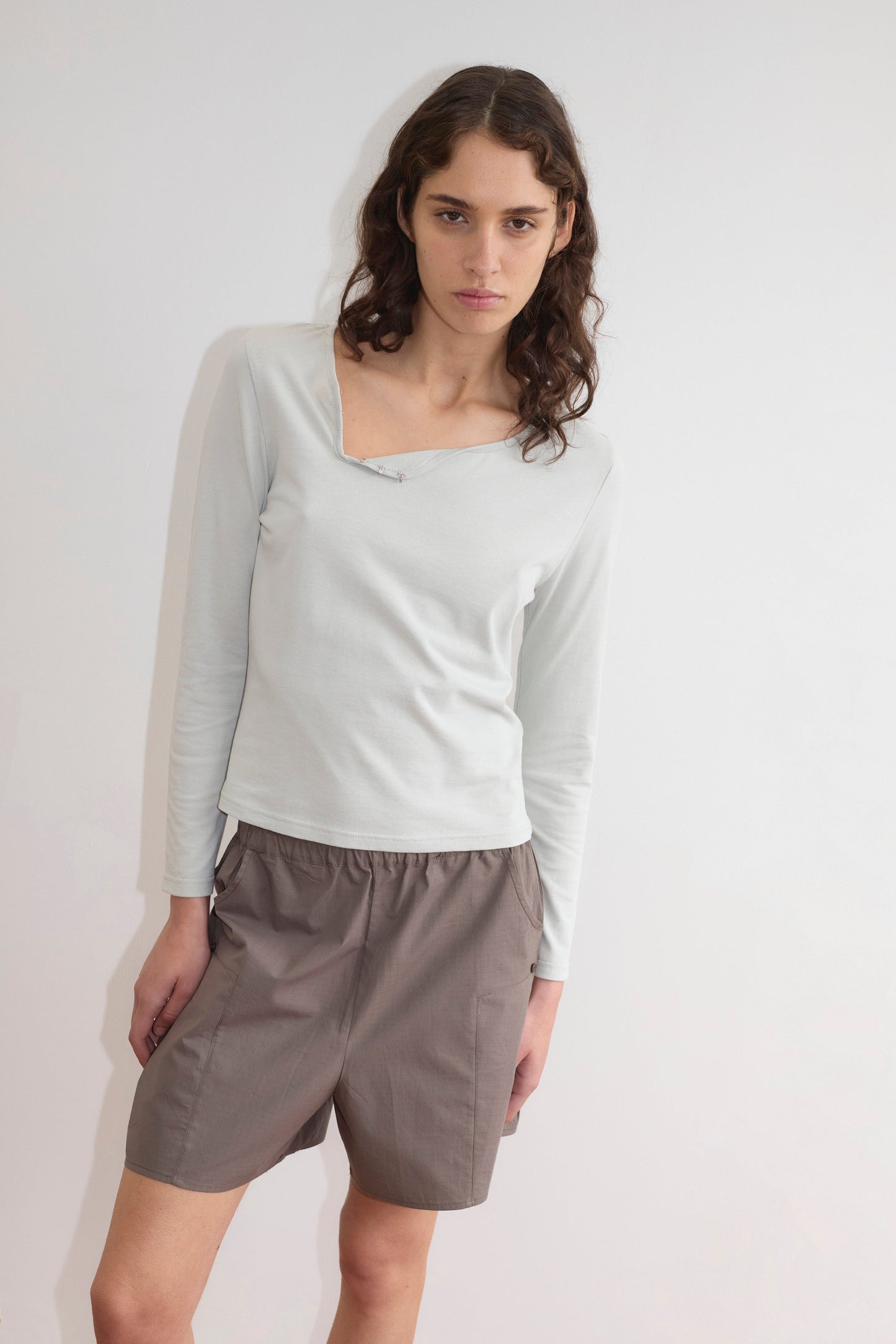 A person with long, wavy brown hair stands against a white wall wearing a Deiji Studios light long-sleeve top with an asymmetrical neckline and Pleat Short - Khaki Check, looking directly at the camera with a neutral expression.
