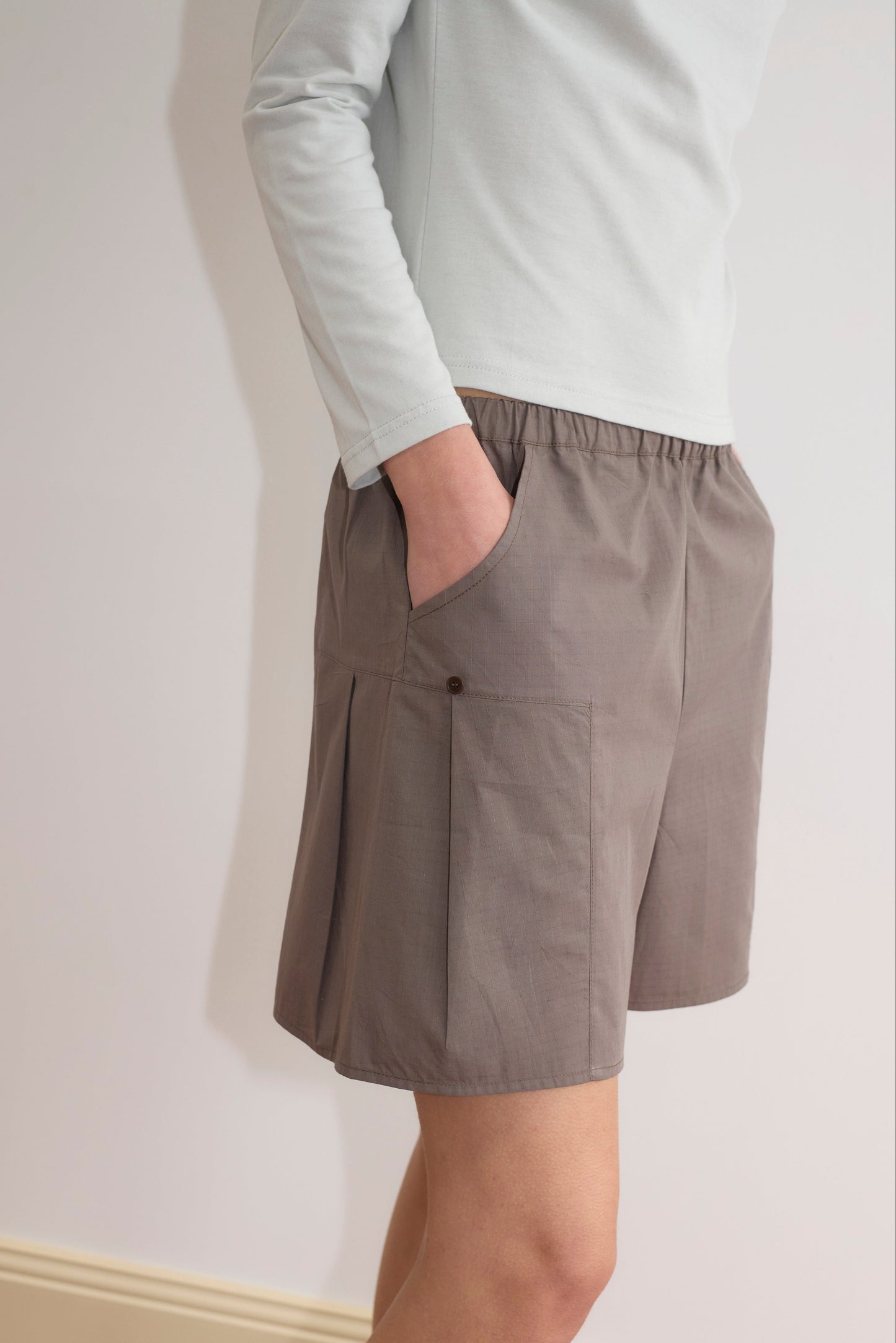 A person wears a light long-sleeve shirt and Pleat Short - Khaki Check with pockets, standing against a plain wall with one hand in their pocket. The outfit looks casual and comfortable.