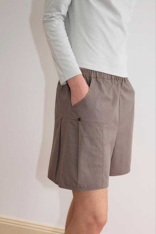 A person wears a light long-sleeve shirt and Pleat Short - Khaki Check with pockets, standing against a plain wall with one hand in their pocket. The outfit looks casual and comfortable.