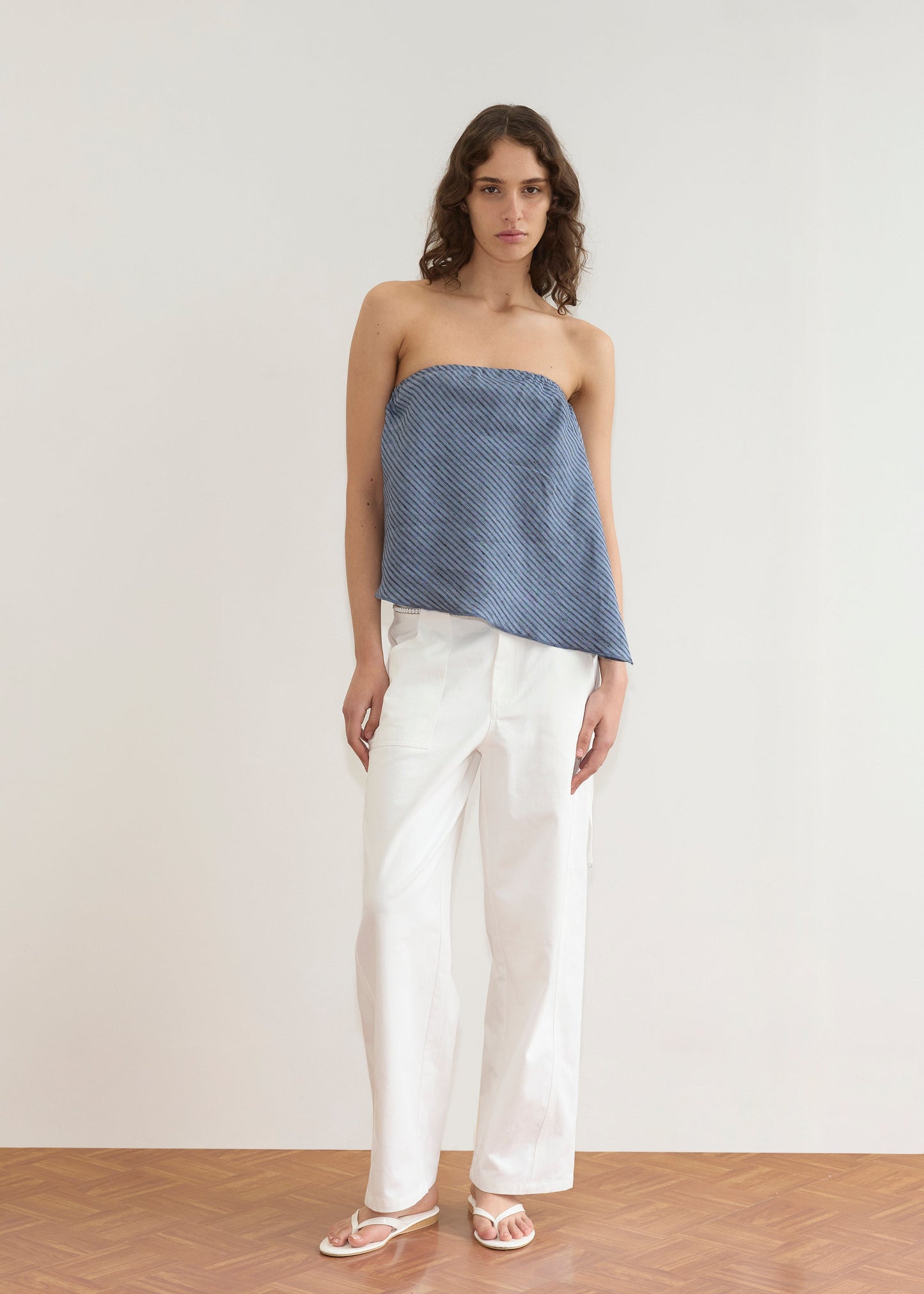 A person with wavy brown hair stands indoors against a light wall, wearing the Line Top - Azure Stripe (a strapless blue top with asymmetric hem), loose white pants, and white sandals on a wooden floor.