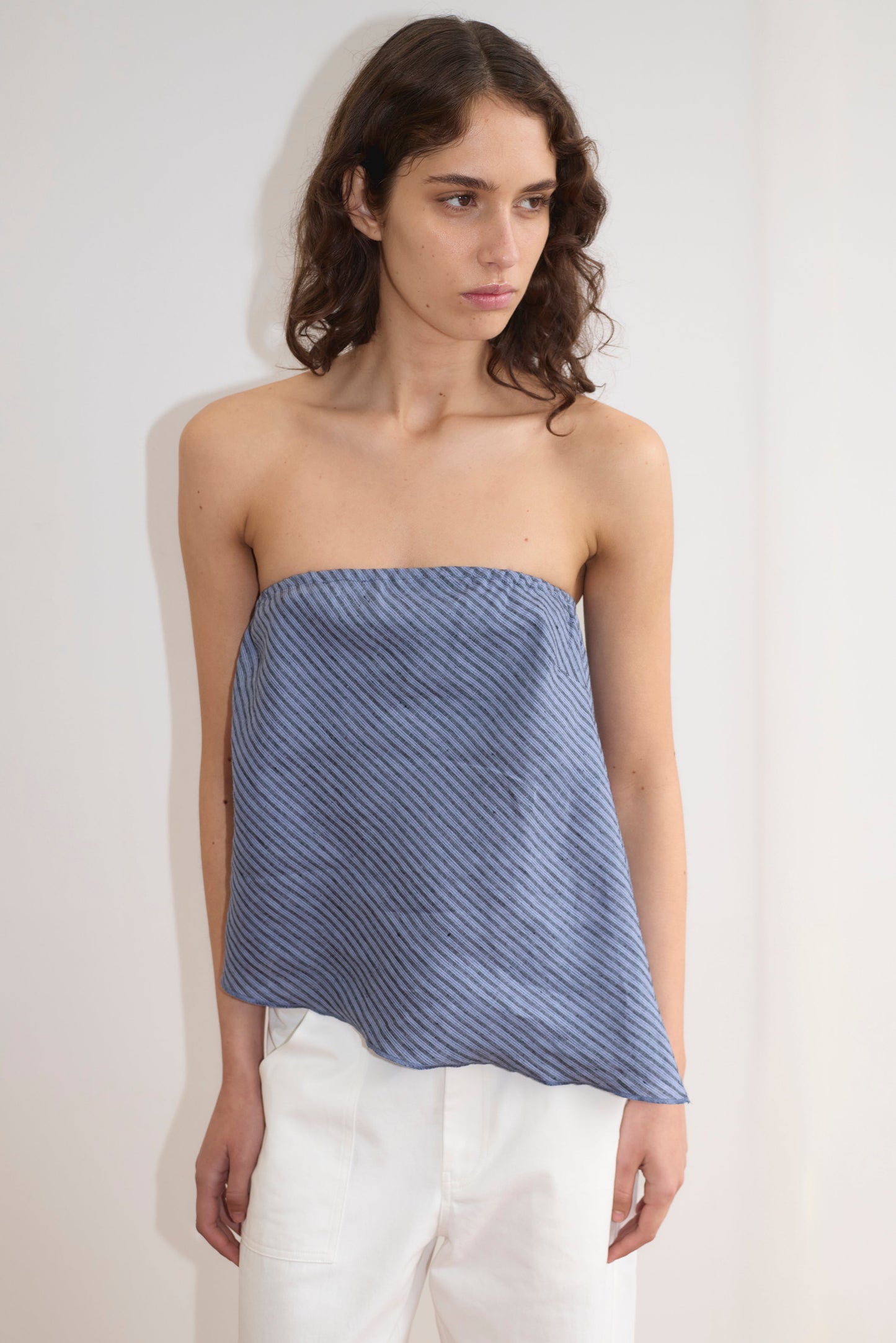 A woman with wavy brown hair wears the Line Top - Azure Stripe, a strapless, lightweight top with diagonal stripes and an asymmetric hem, paired with high-waisted white pants against a plain light background.