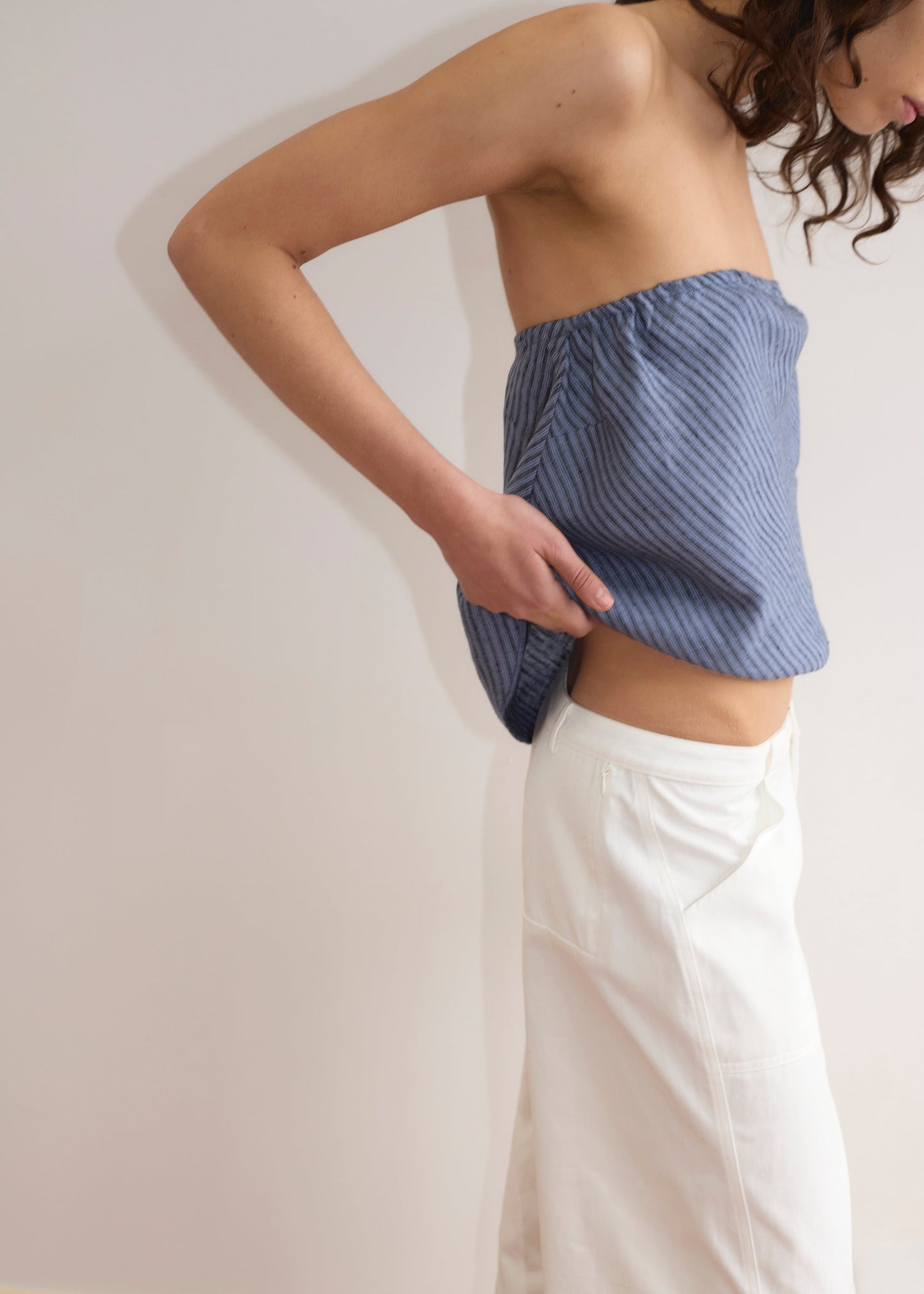 A person wearing the Line Top - Azure Stripe, a blue strapless top with an asymmetric hem, and white pants stands sideways against a light background, adjusting the top with one hand while their face is partially out of frame.