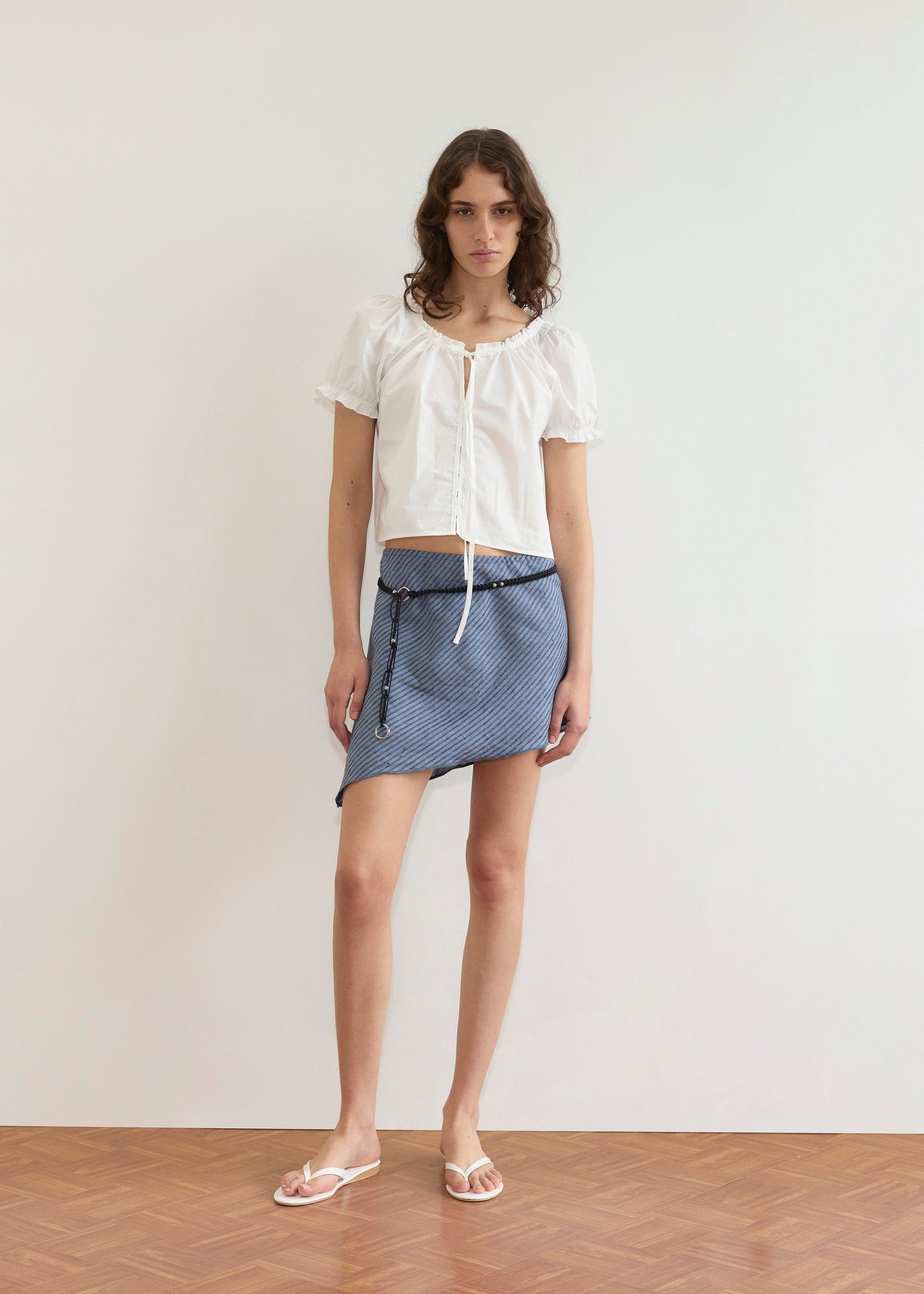 A person with wavy hair stands indoors on a wooden floor against a plain white wall, wearing the Close Shirt - White (short sleeves, cropped relaxed fit), a blue and white striped asymmetrical mini skirt, and white flip-flops.
