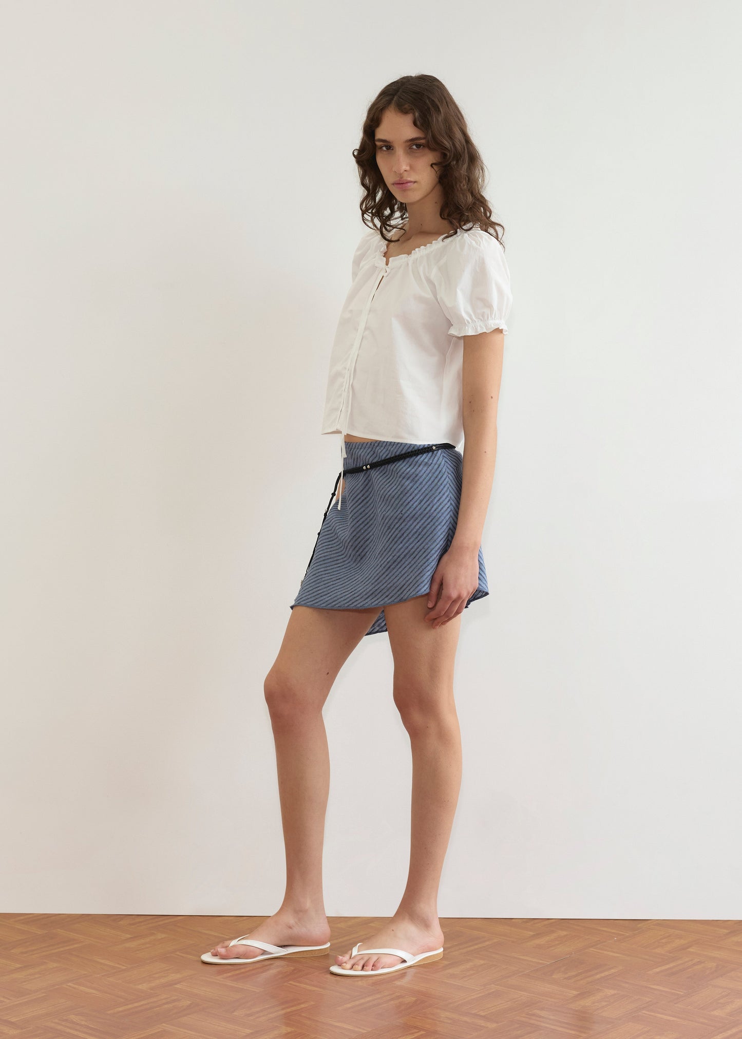 A young woman with wavy brown hair stands indoors wearing the Close Shirt - White, a blue patterned mini skirt, and white sandals. She looks slightly toward the camera, posed against a plain background.