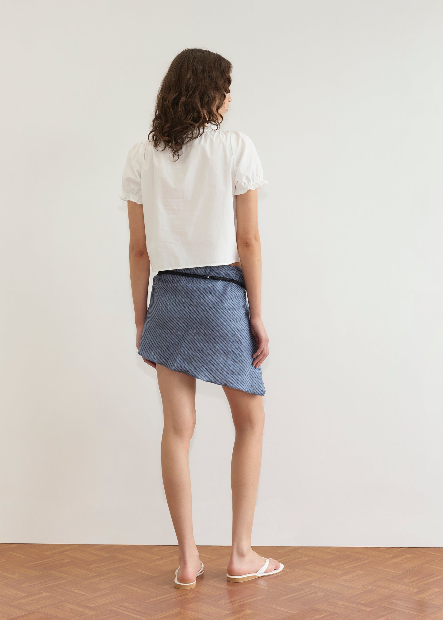 A woman stands facing away, wearing the Close Shirt - White with short sleeves and a cropped relaxed fit, paired with a blue pinstriped asymmetrical skirt and white sandals on a wooden floor against a plain white wall.