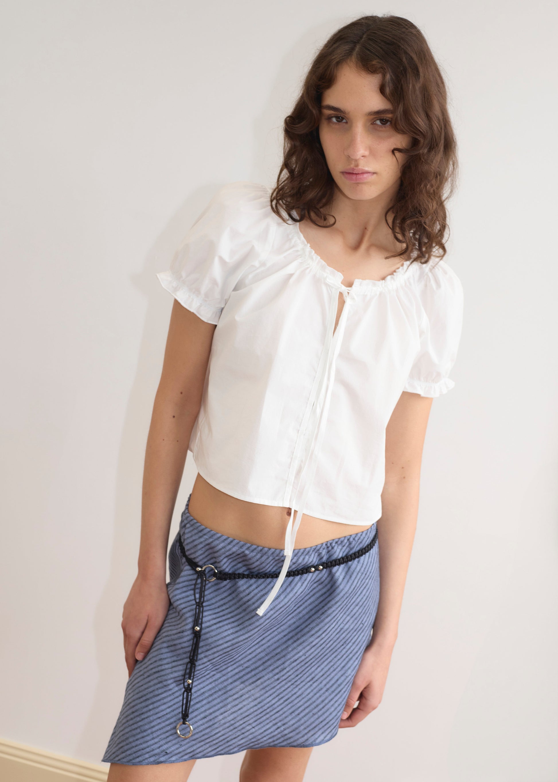 A woman with wavy brown hair wears the Line Top - Azure Stripe—a lightweight, white short-sleeve blouse with a tie neckline—paired with a blue striped skirt and black belt, standing against a plain white wall.
